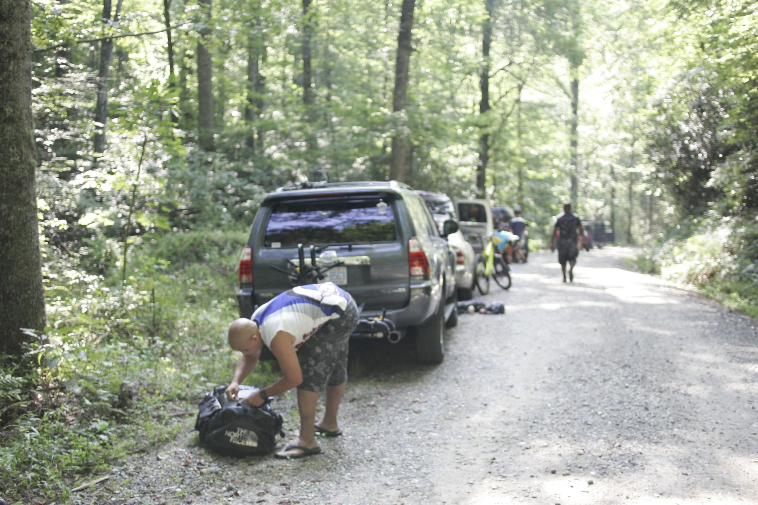 A man is bending down to open a backpack beside parked vehicles on a gravel road surrounded by lush greenery. Another person is walking in the background, and several bicycles are visible near the cars. Sunlight filters through the trees, creating a bright and scenic outdoor atmosphere. Laurel Mountain mountain bike trail.