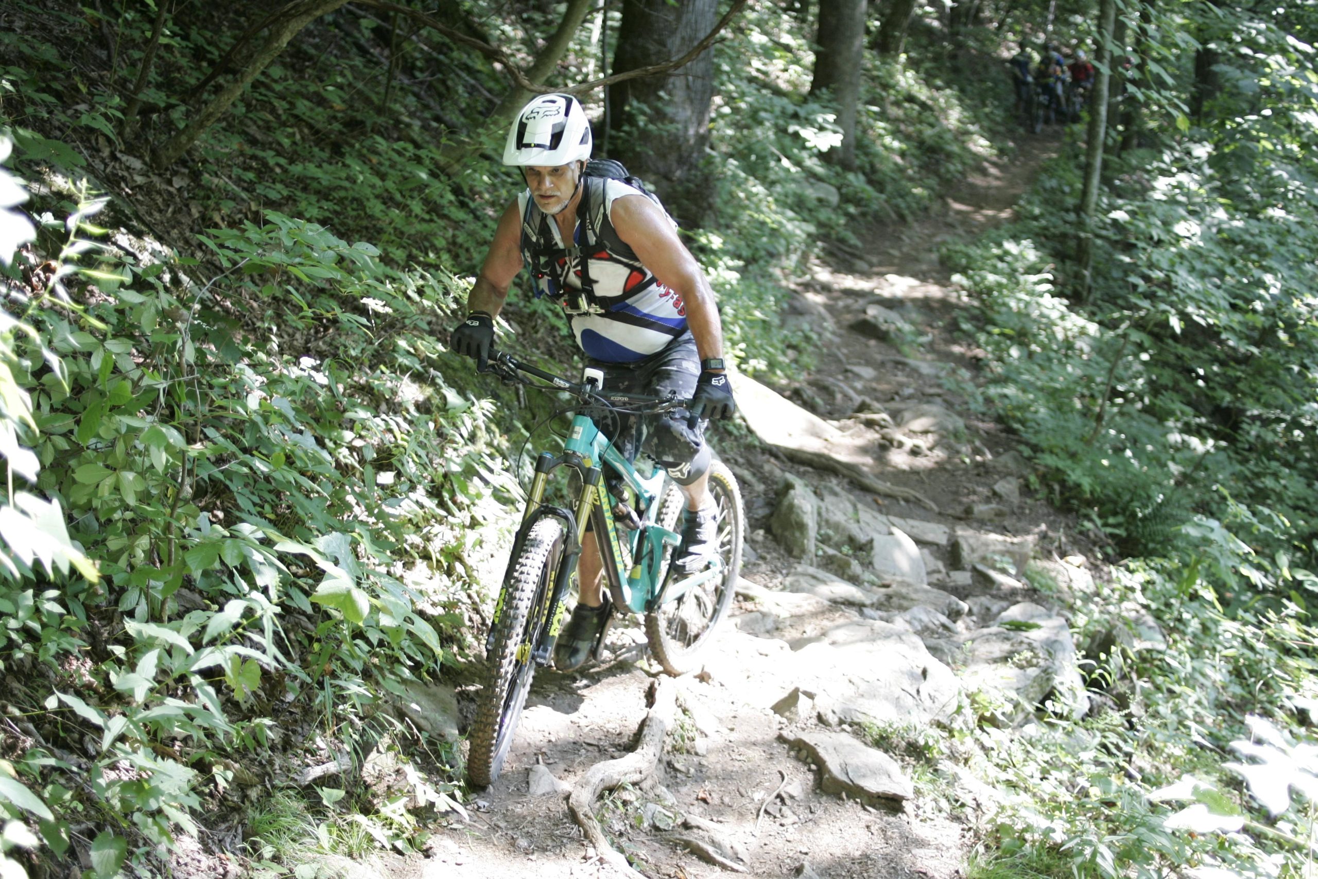 A mountain biker navigating a rocky, narrow trail surrounded by dense greenery and trees, wearing a helmet and protective gear. The biker appears focused and is maneuvering their bike over the challenging terrain. Laurel Mountain mountain bike trail.