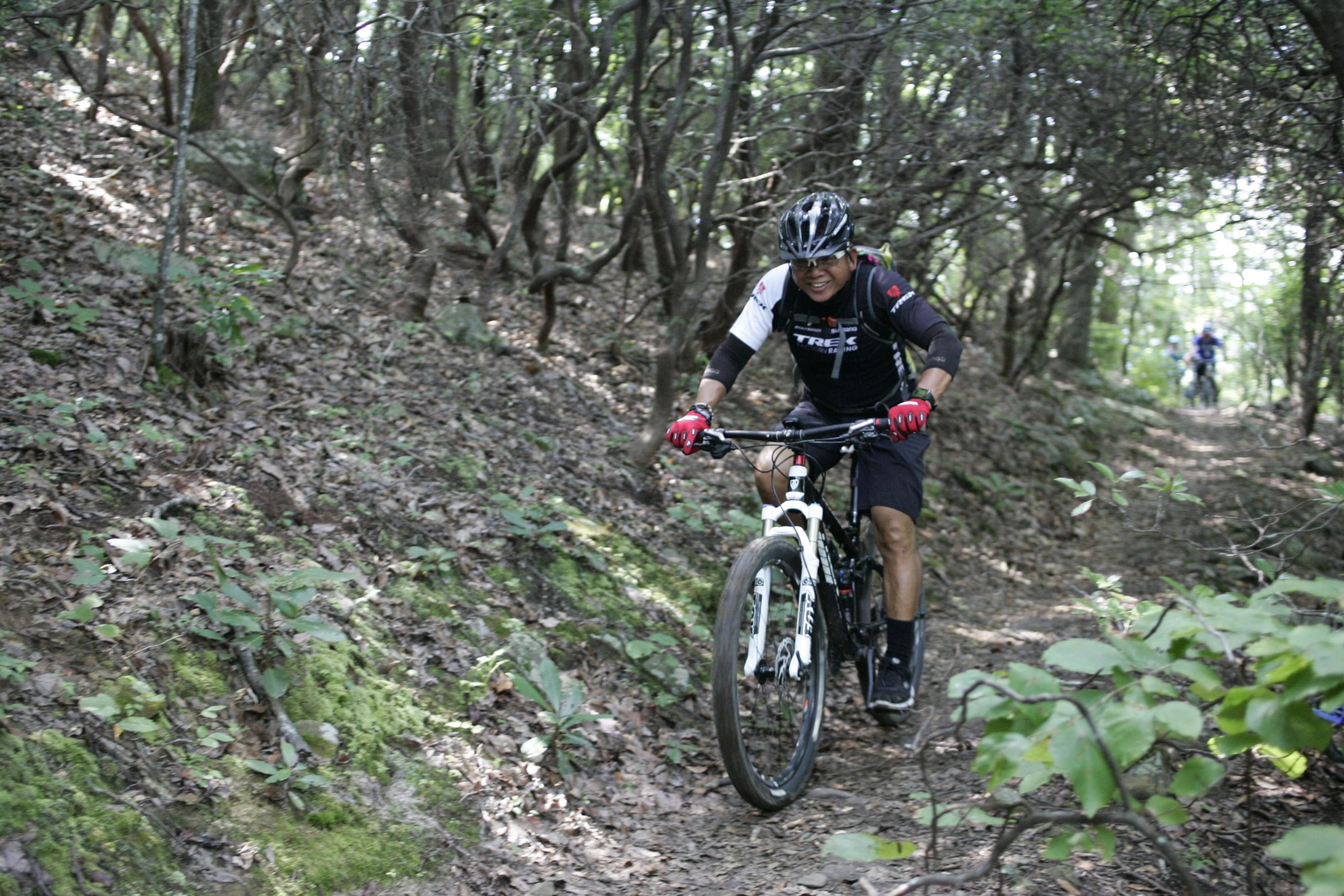A cyclist navigating a rocky, wooded trail on a mountain bike, focused on maintaining balance. The terrain is covered with leaves and greenery, and the sunlight filters through the trees. Another cyclist can be seen in the background. Laurel Mountain mountain bike trail.