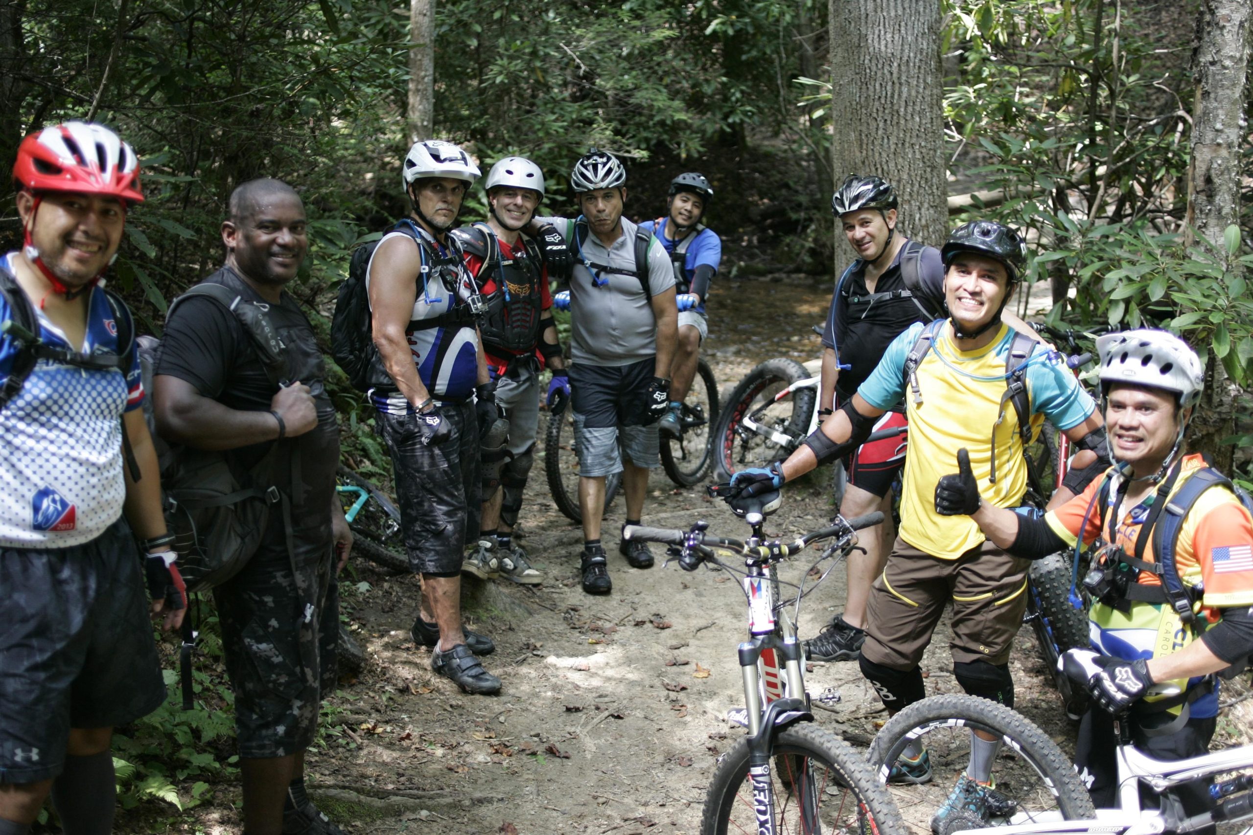 A group of eight mountain bikers is posing on a dirt trail surrounded by lush greenery. They are wearing helmets and protective gear, and several bikes are leaning against the trail. The riders, smiling and relaxed, showcase a mix of colorful jerseys and shorts, indicating a lively day of biking in a natural setting. Laurel Mountain mountain bike trail.