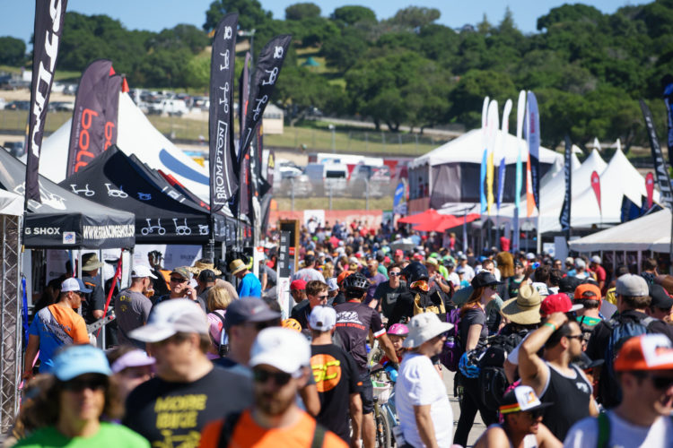 A busy outdoor event with a large crowd walking through vendor tents and booths. Various flags and canopies display bicycle brands, and people of all ages are engaged in activities, some wearing helmets and carrying bikes. The background features trees and a racetrack.
