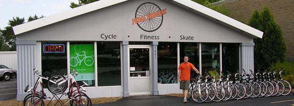 A fitness store exterior with the sign "Open" lit up. In front, a smiling employee stands next to a row of bicycles for sale. The building has a modern design with large windows displaying a bicycle graphic. The signs above the entrance read "Cycle," "Fitness," and "Skate." 