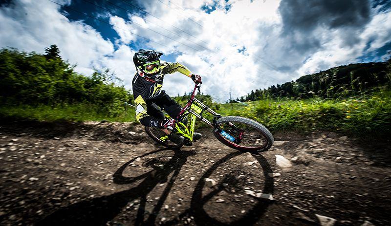 A mountain biker in vibrant gear leans into a turn on a dirt trail, surrounded by lush greenery and a cloudy sky. The image captures dynamic movement and the thrill of downhill biking. The Mini DH mountain bike trail.