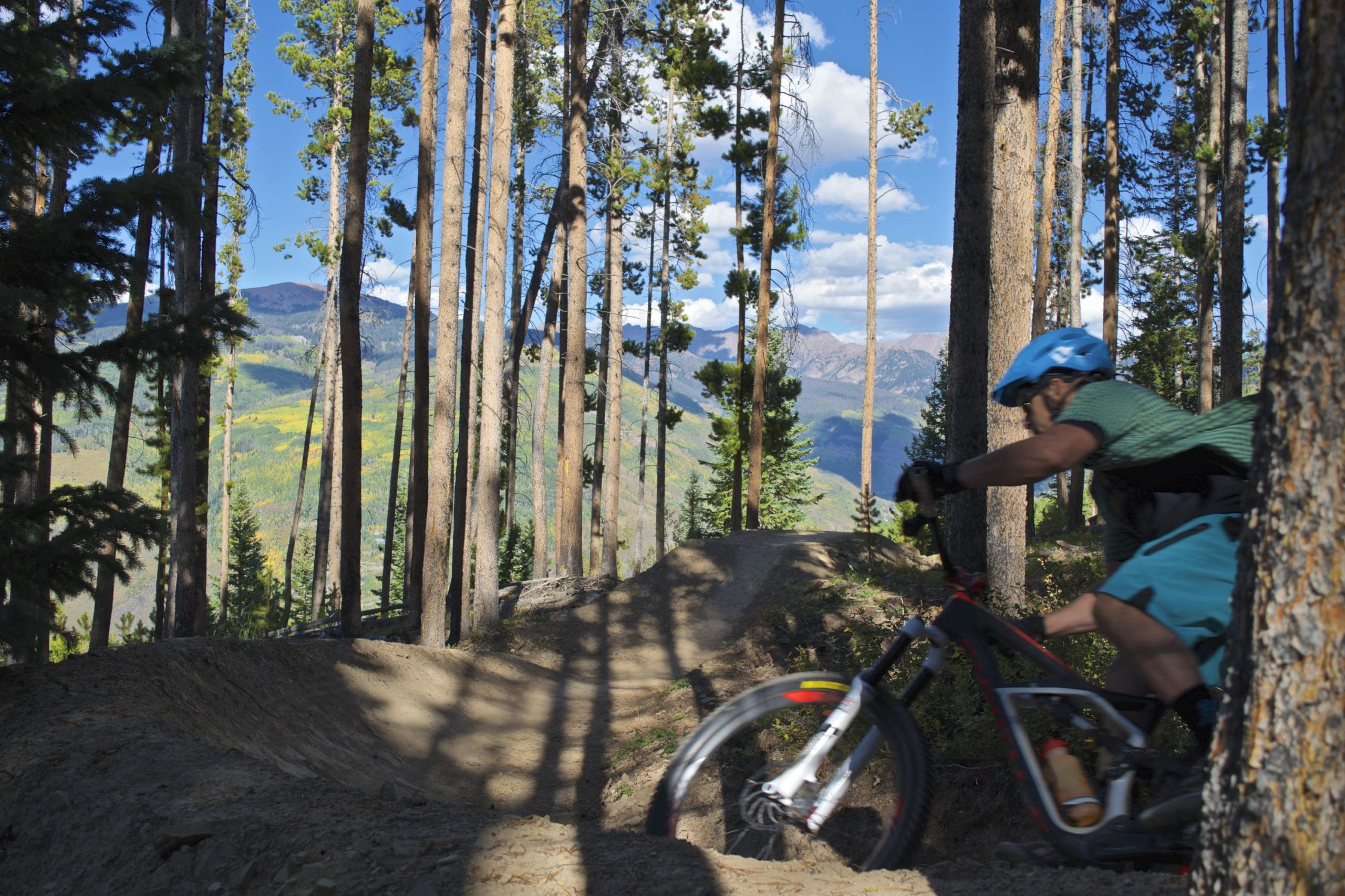 A mountain biker riding along a dirt trail through a forest of tall pine trees, with mountains and a blue sky in the background. The biker is captured mid-action, leaning into a turn, wearing a blue helmet and a green shirt. The scene showcases vibrant green and yellow foliage on the hillside, indicating a beautiful outdoor environment. Vail Mountain Bike Park mountain bike trail.