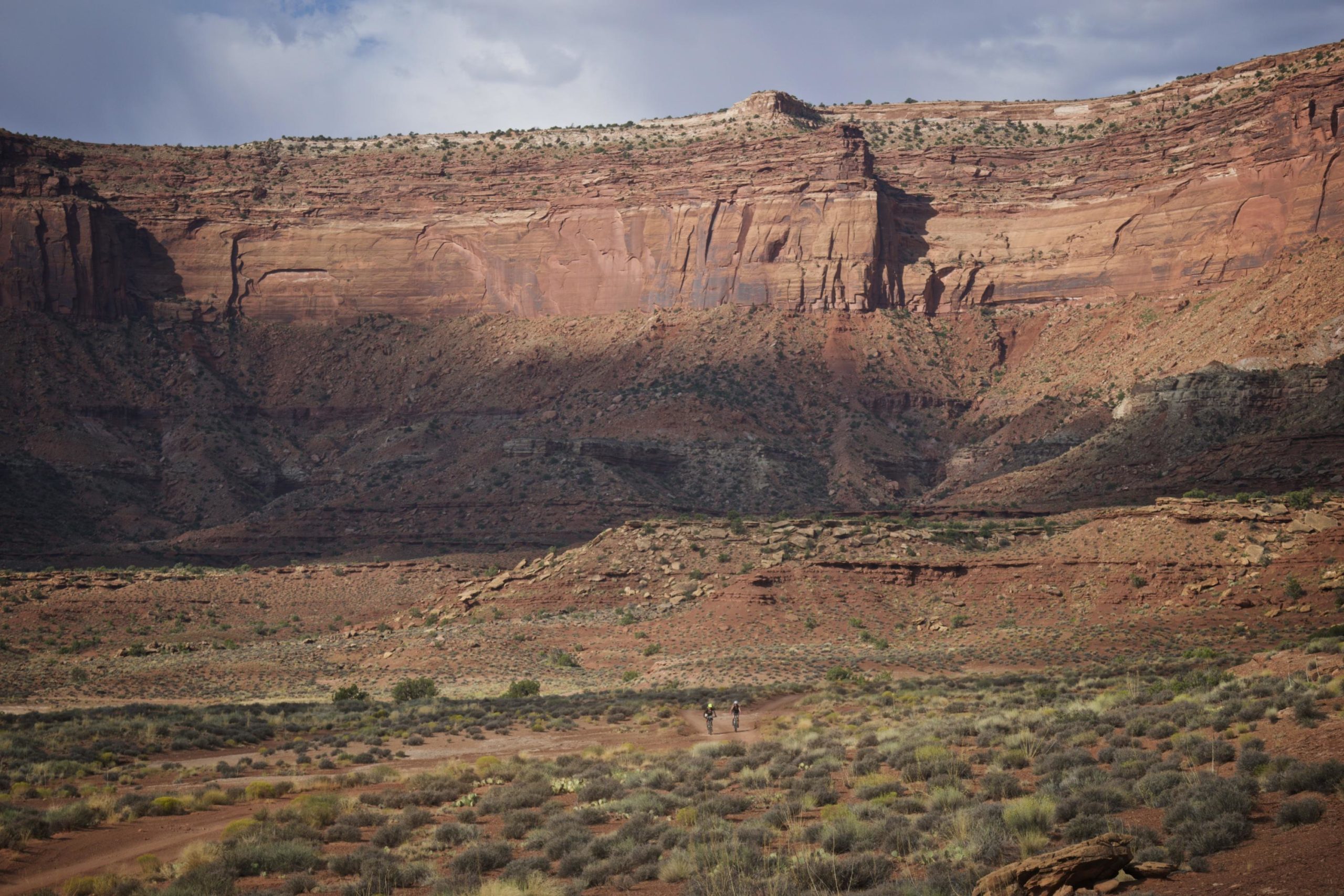 A scenic desert landscape featuring towering red rock formations and a rugged terrain. In the foreground, two people are walking along a dirt trail, surrounded by sparse vegetation and scattered shrubs. The dramatic cliffs rise in the background under a cloudy sky. White Rim Trail mountain bike trail.