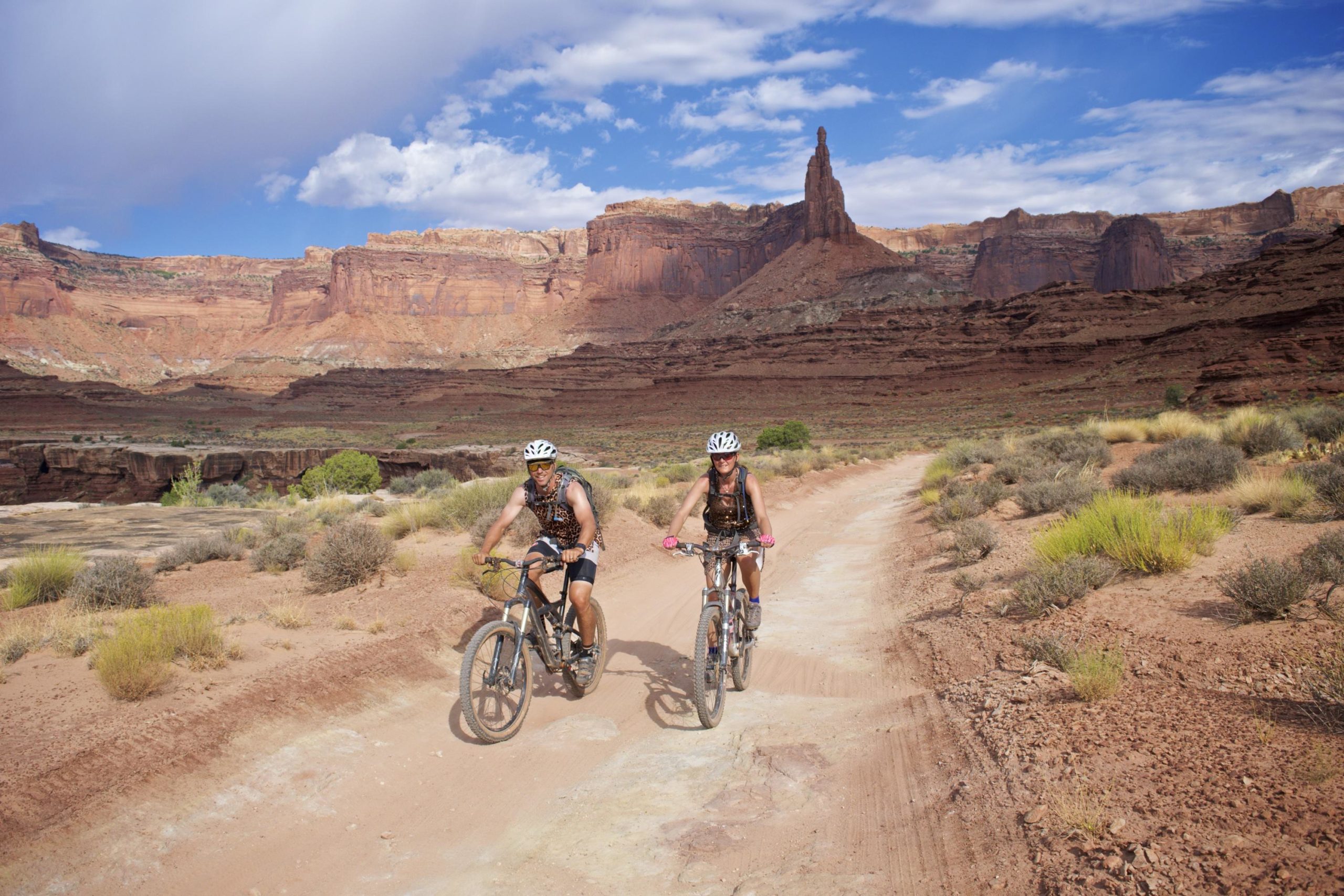 Two mountain bikers ride along a dirt trail in a rugged landscape featuring towering red rock formations and a partly cloudy sky. The area is sparse with vegetation, showcasing the natural beauty of the desert terrain. White Rim Trail mountain bike trail.