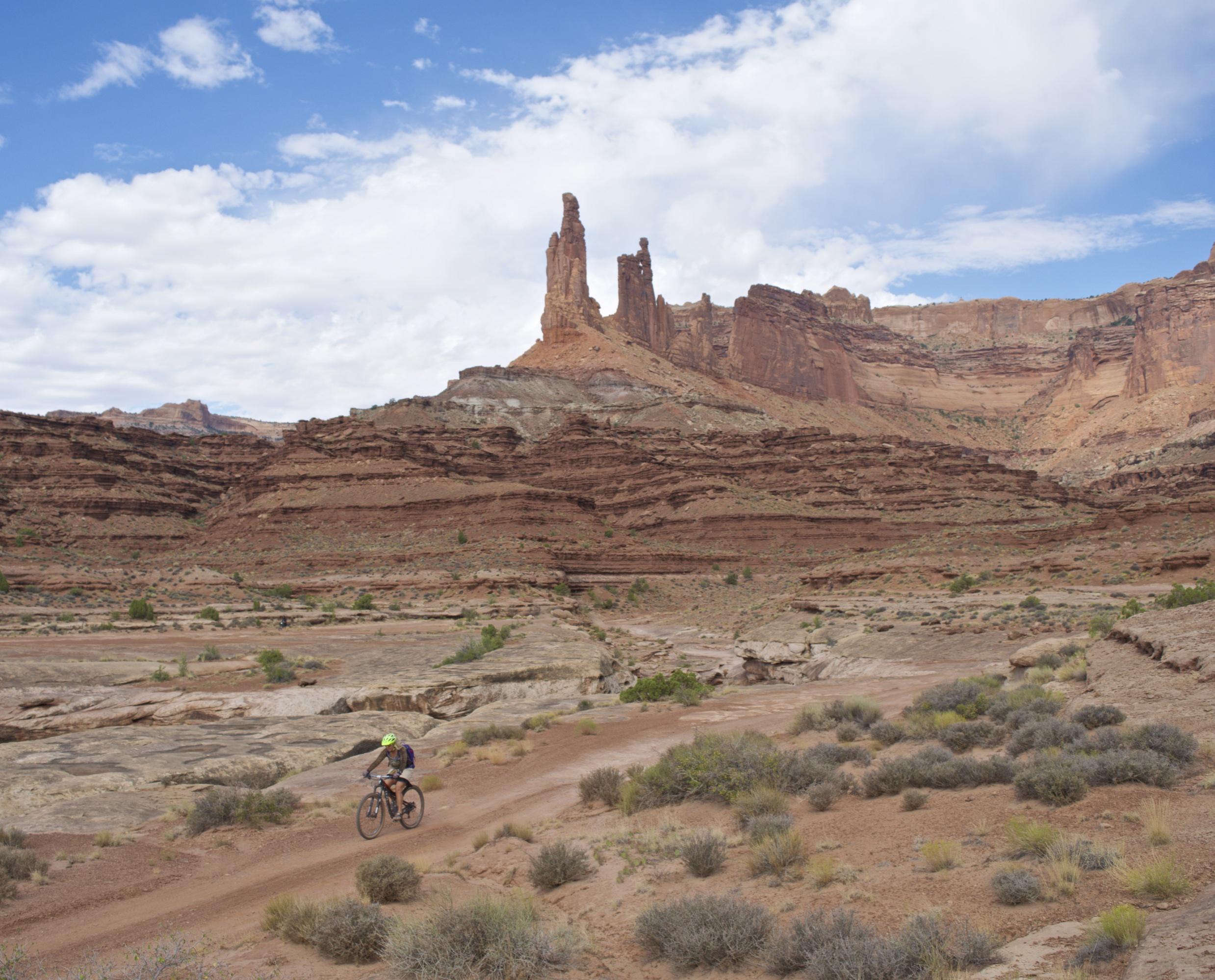 A mountain biker rides along a dirt trail in a rugged desert landscape featuring towering red rock formations and a partly cloudy blue sky. The terrain is characterized by layers of rock and sparse vegetation, with the biker navigating through the natural beauty of the outdoors. White Rim Trail mountain bike trail.