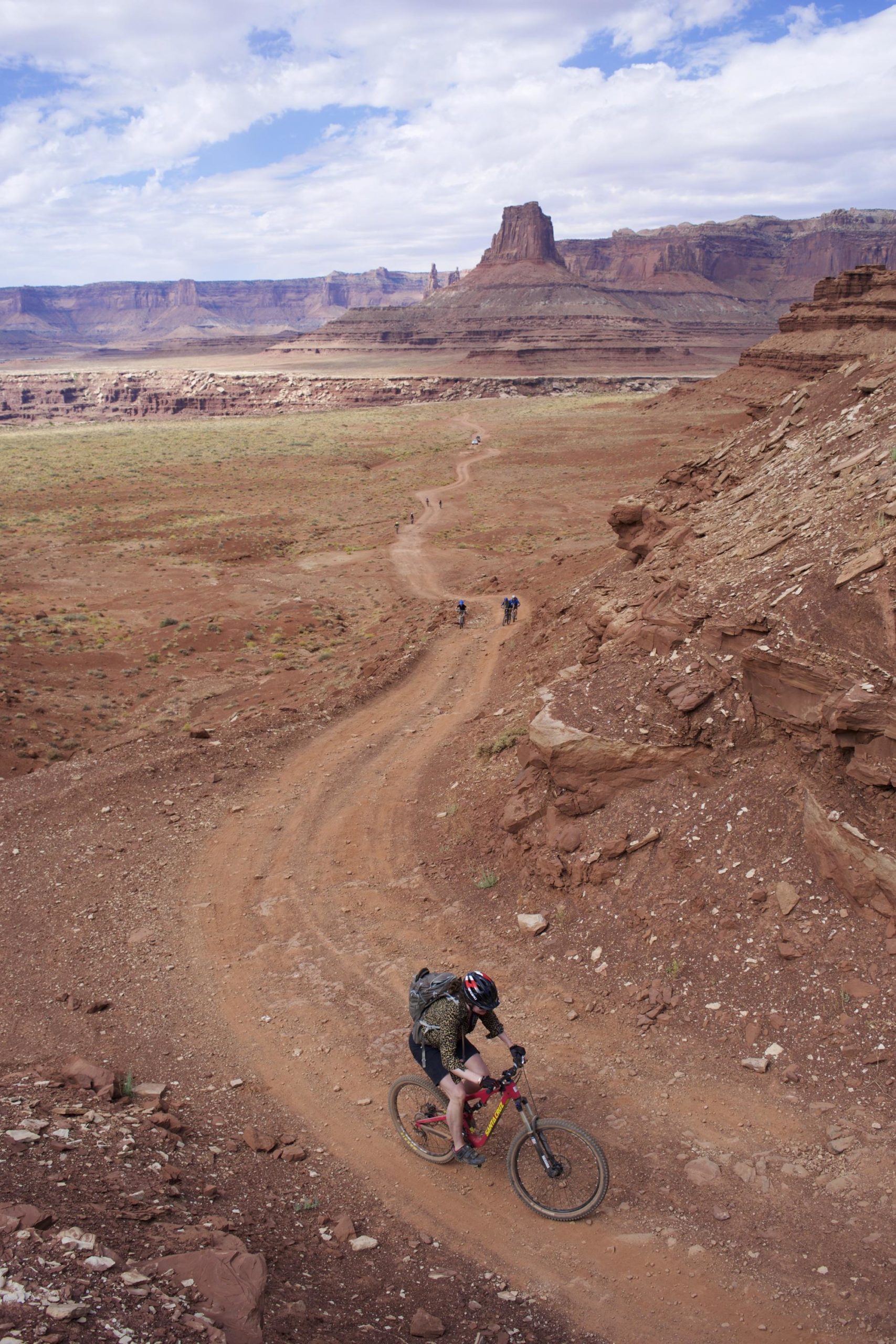 A cyclist navigates a winding dirt trail through a rugged, arid landscape, with distant rock formations and cloudy skies in the background. Several other hikers can be seen on the trail, adding to the adventurous atmosphere of the scene. White Rim Trail mountain bike trail.