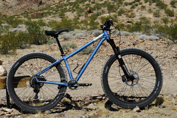 A blue mountain bike resting on a rocky terrain with sparse vegetation in the background. The bike features wide tires, a front suspension fork, and a prominent gear system. The landscape is arid, suggesting a rugged outdoor environment suitable for biking.
