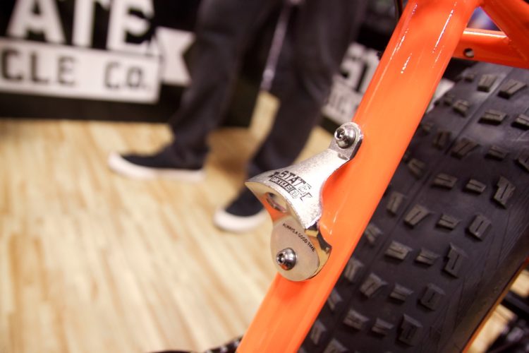 Close-up of an orange bicycle frame featuring a silver bell attached, with a focus on the bell's detailing. The background shows a portion of a logo and a blurred figure wearing dark clothing, indicating a setting likely at a bicycle event or exhibition.