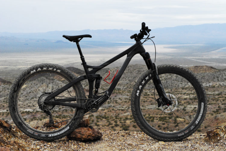 A black mountain bike is positioned on rocky terrain, overlooking a vast desert landscape with mountains in the distance. The bike features wide tires, a sturdy frame, and a suspension system, emphasizing its off-road capabilities. The sky is overcast, creating a dramatic backdrop for the scene.