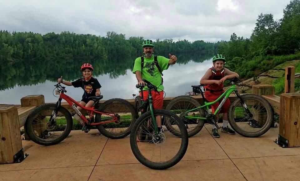 Surly Krampus: A man and two boys stand on a deck by a calm lake, each holding their mountain bikes. The man wears a bright green shirt and is giving a thumbs-up, while the boys are smiling and posing with their bikes—one is red and the other green. The background features lush greenery and a cloudy sky reflecting on the water's surface.