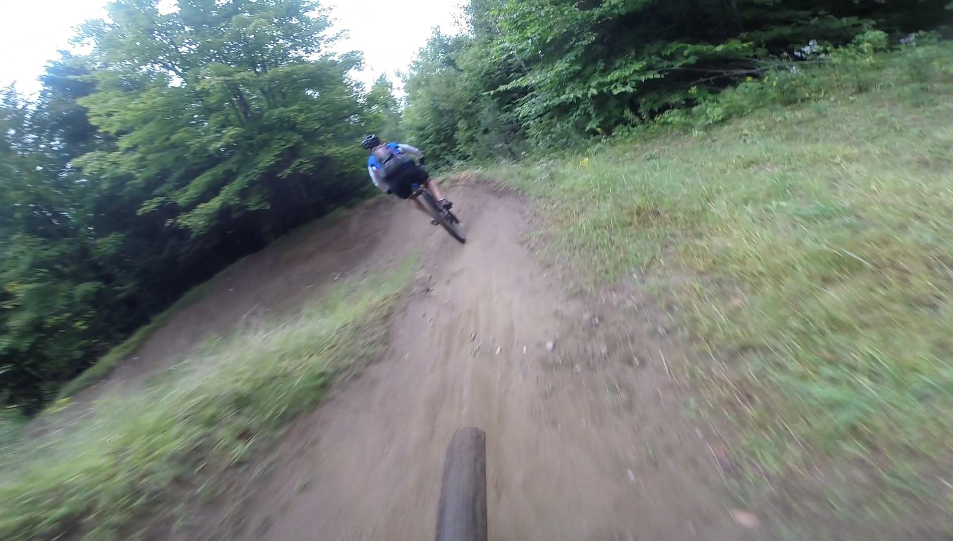 A mountain biker navigating a dirt trail surrounded by trees, captured from a low-angle perspective showing the bike's front tire and the rider in motion as they take a curve on the path. Kingdom Trails mountain bike trail.