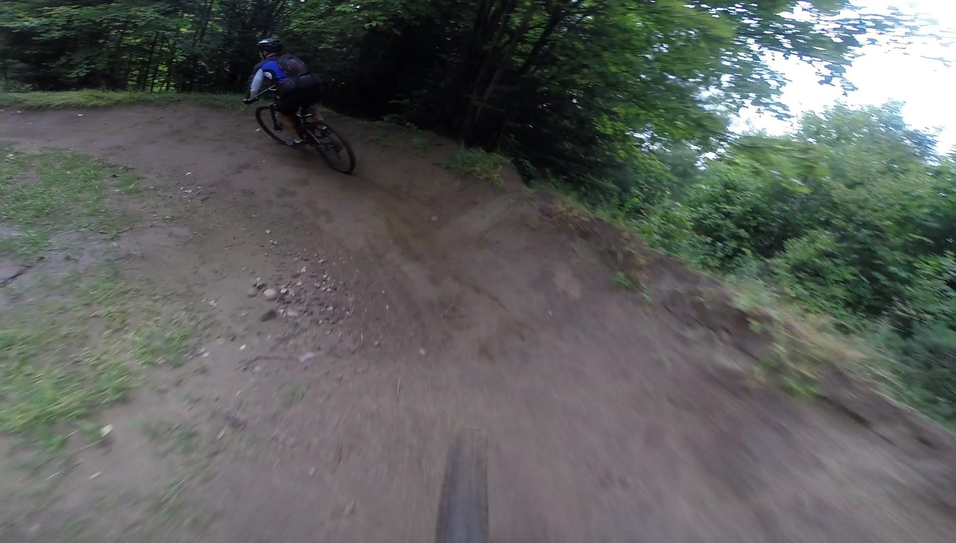 A mountain biker navigates a dirt trail with a slight curve, surrounded by lush green foliage and trees. The cyclist is in motion, wearing a helmet and a backpack, while the terrain shows signs of recent activity. Kingdom Trails mountain bike trail.
