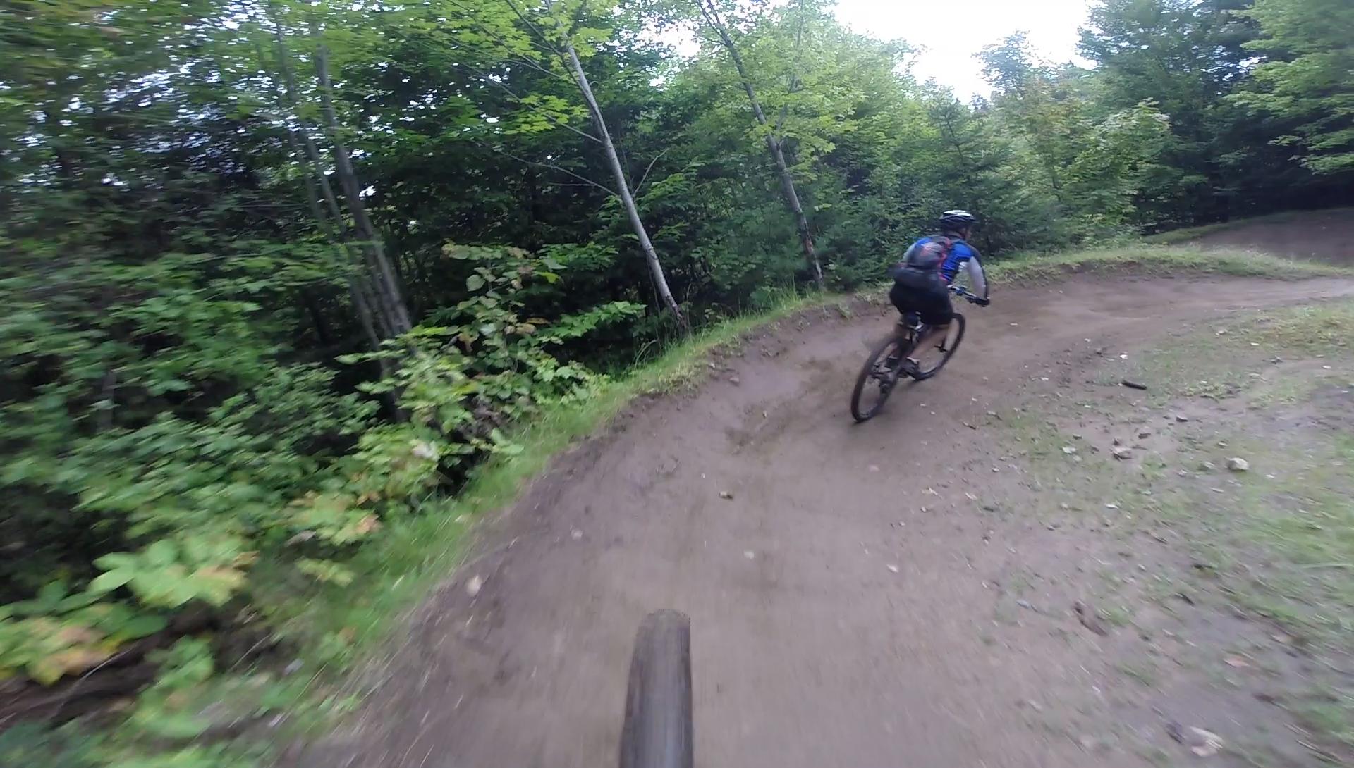 A mountain biker navigating a curved dirt trail surrounded by lush green trees and underbrush. The perspective shows part of the biker's wheel in the foreground, emphasizing speed and motion through the natural environment. Kingdom Trails mountain bike trail.
