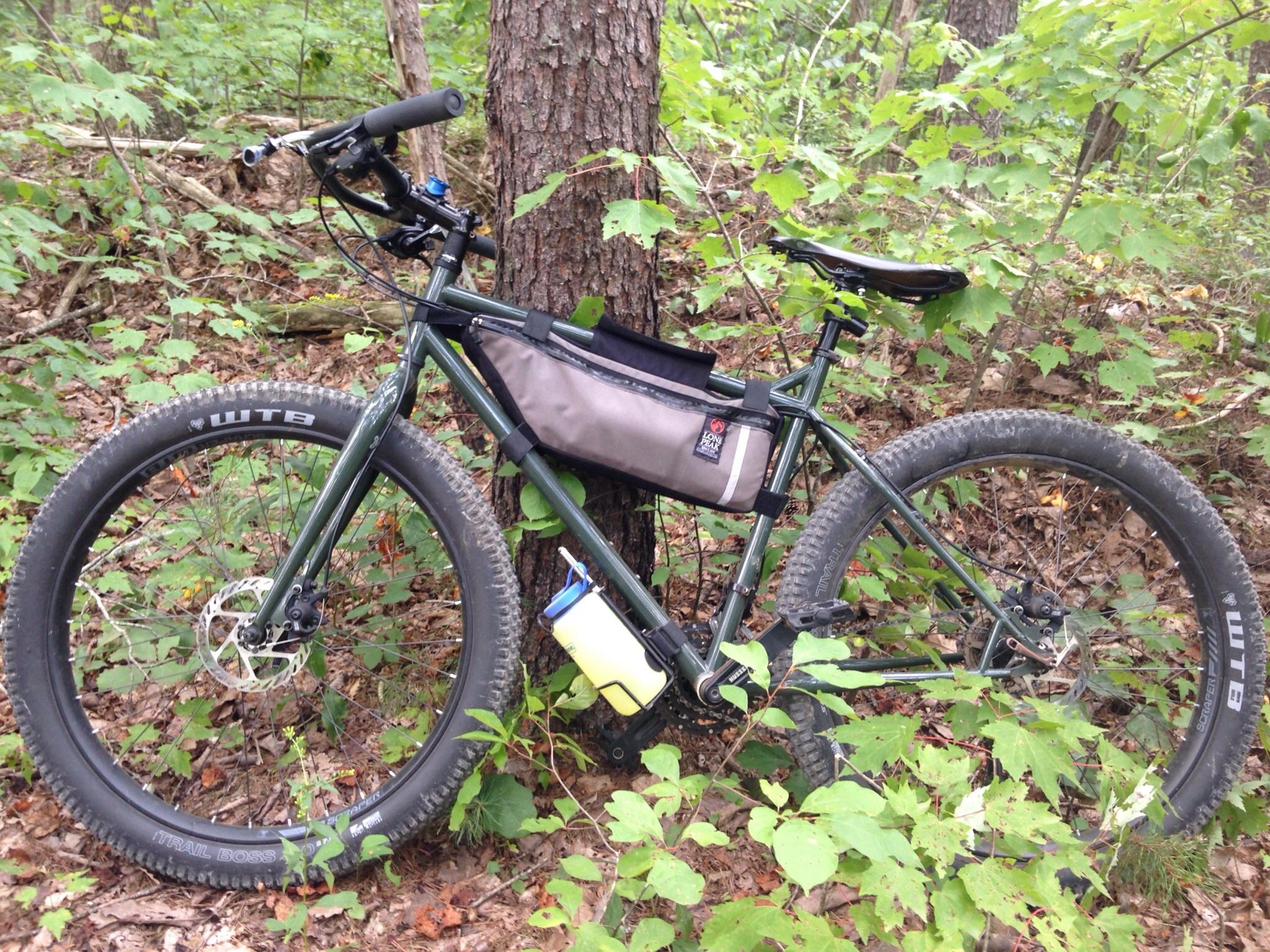 Nashbar Big Ol Fat Bike: A mountain bike resting against a tree in a forested area, surrounded by green foliage and underbrush. The bike features thick tires and a brown bag mounted on the frame for carrying supplies.