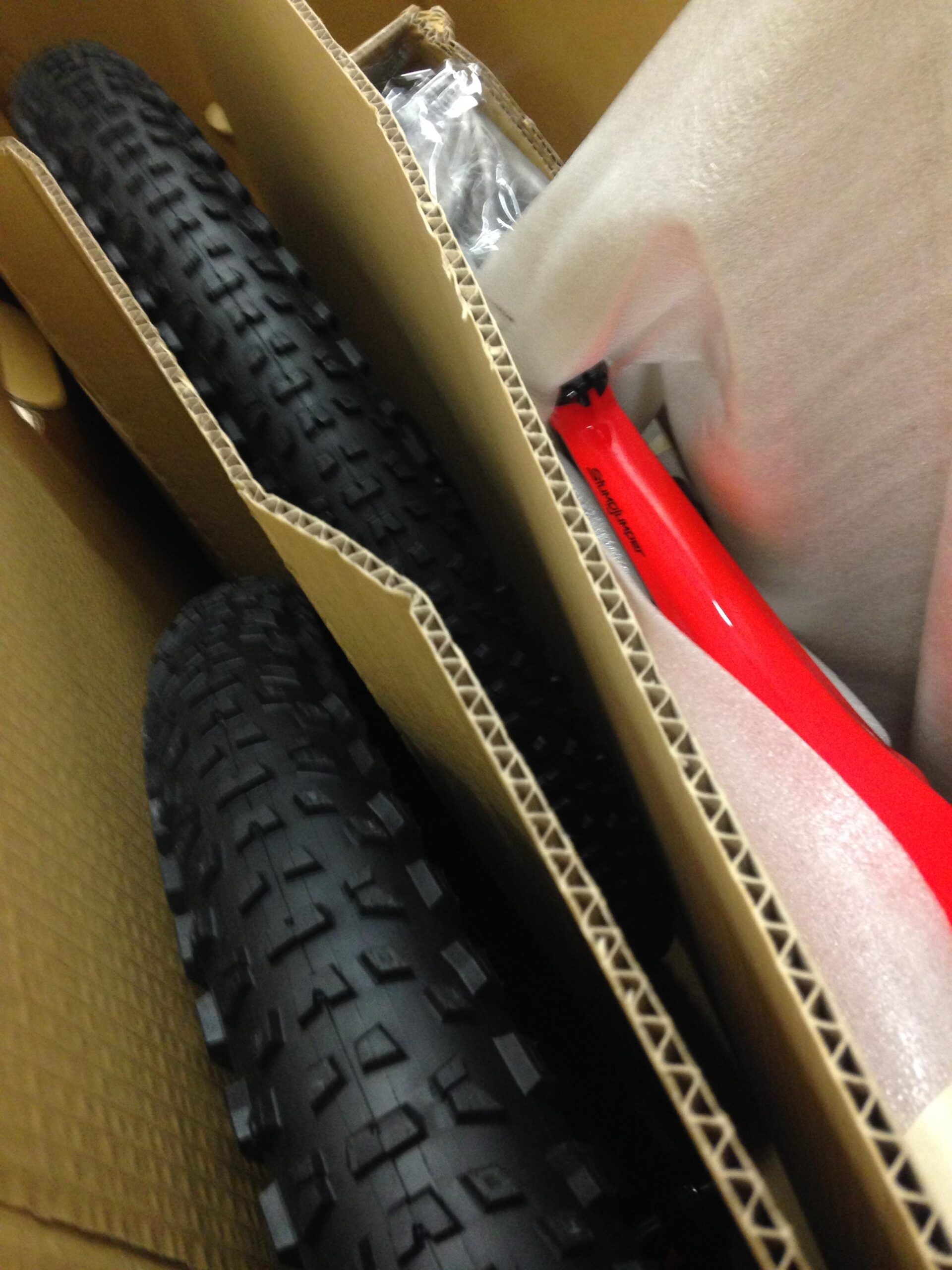 A close-up view of boxed bicycle parts, including two black tires with textured surfaces and a bright red bike frame partially wrapped in protective material, all arranged in a cardboard box.