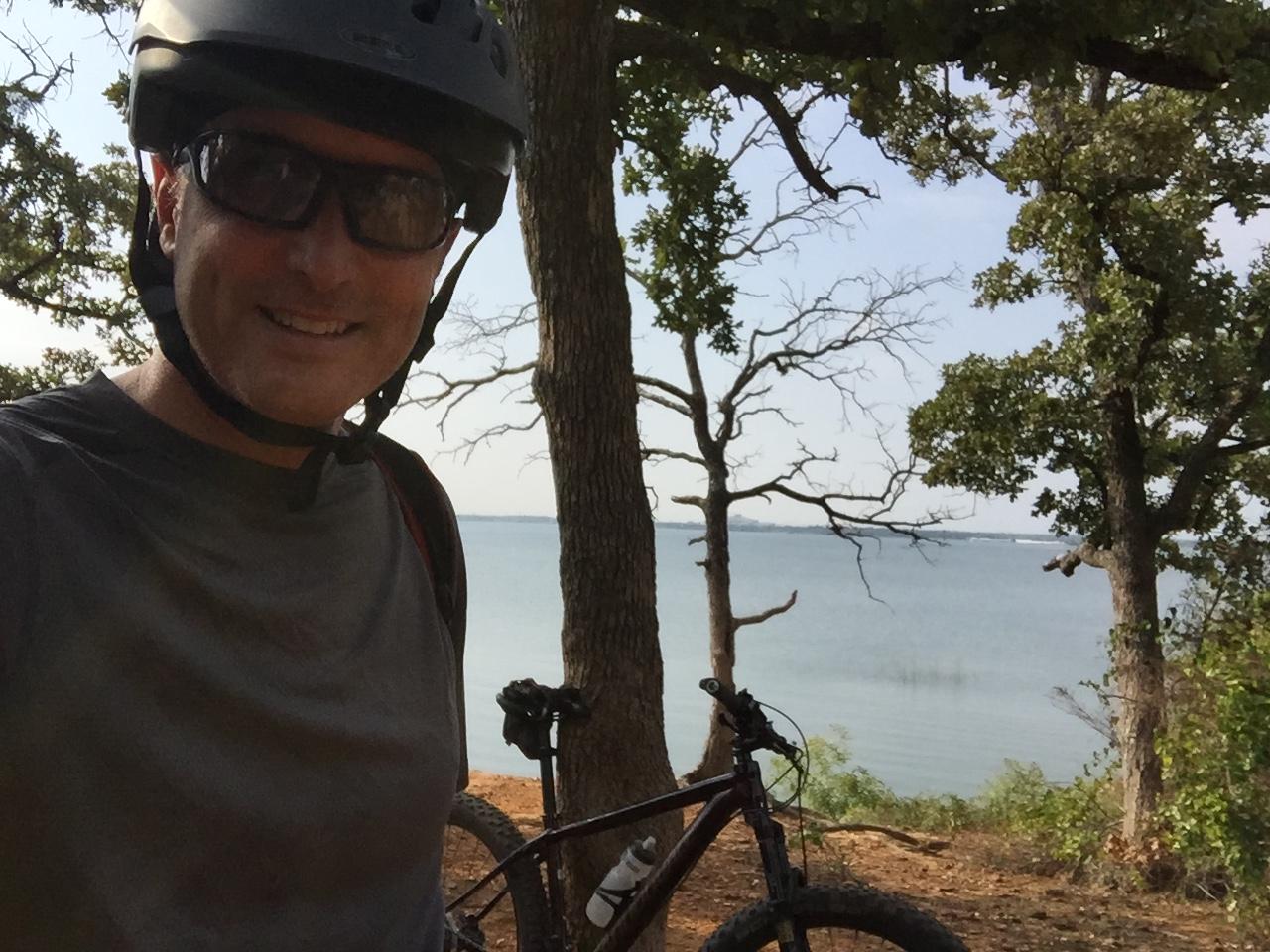 A person wearing a bicycle helmet and sunglasses smiles at the camera while standing near a mountain bike. The background features trees and a body of water, suggesting a scenic outdoor location. Northshore Trail mountain bike trail.
