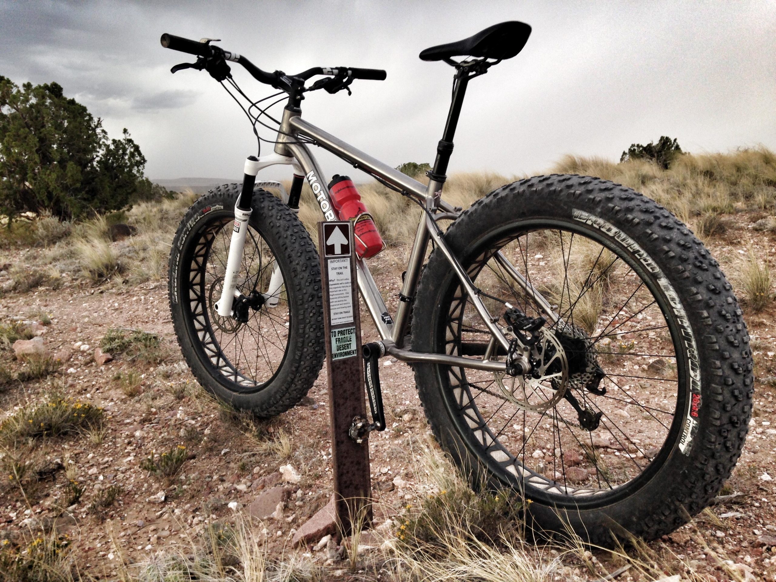 A fat bike with large tires is parked next to a sign in a rugged outdoor setting. The bike's frame is silver with black handlebars, and it has two water bottles mounted on the frame. Behind the bike, there are sparse grasses and shrubs under a cloudy sky. The sign nearby notes important information about protecting the fragile desert environment. Mariposa Fat Bike Trails mountain bike trail.