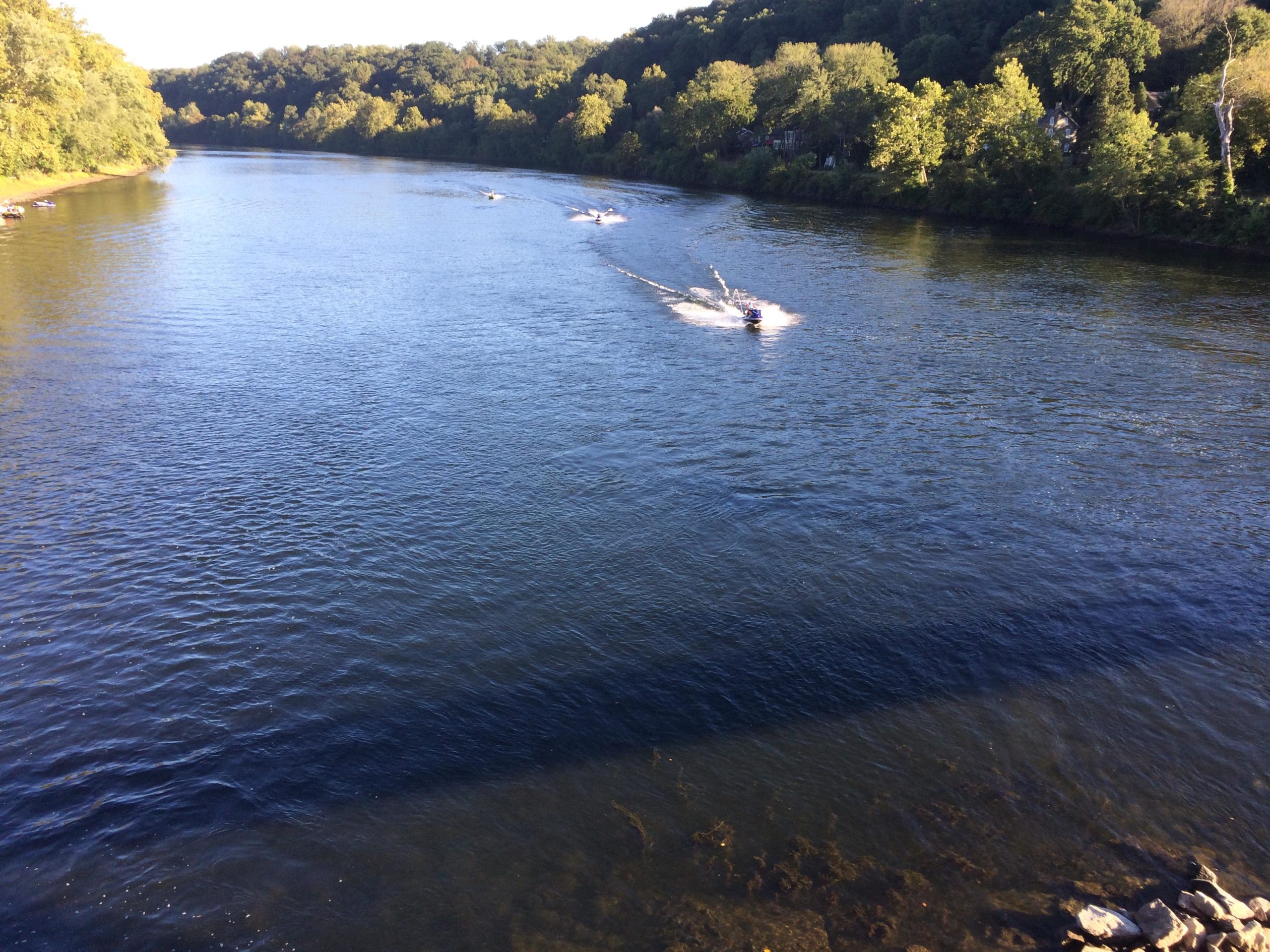 A scenic view of a calm river surrounded by trees, with two boats racing on the water's surface. The sun casts a warm light, creating reflections on the water, while the riverbanks are lined with greenery. Bridge To Bridge - D&r Canal mountain bike trail.