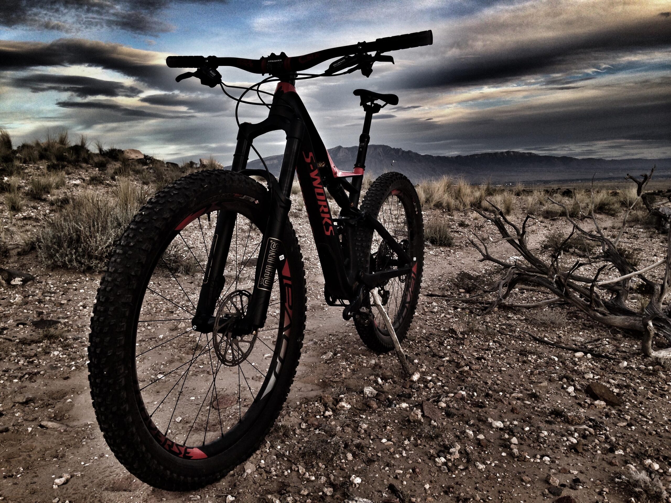 Specialized Stumpjumper FSR 6Fattie: A black mountain bike with distinctive red accents is positioned on a gravel path, surrounded by sparse vegetation and rocky terrain. The backdrop features an expansive sky with dramatic clouds and distant mountains, creating a rugged outdoor setting.
