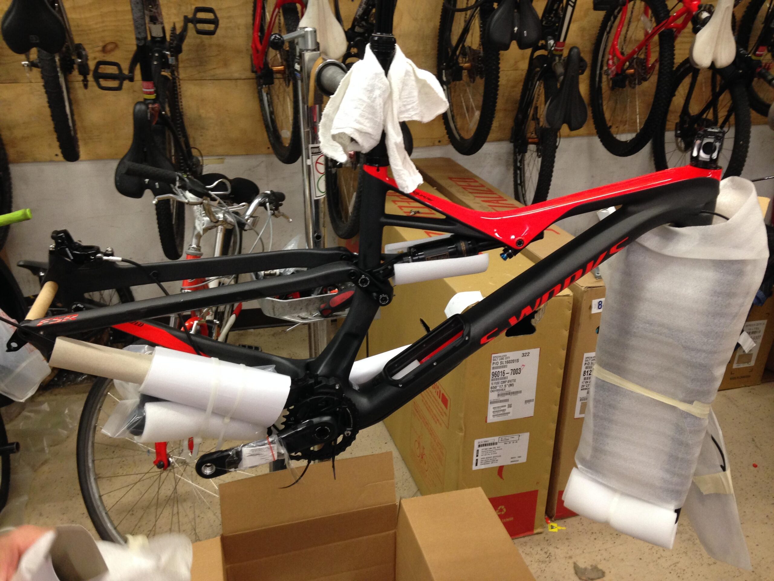 A black and red bicycle frame is partially assembled and wrapped in protective materials, sitting among boxes and other bikes in a workshop setting. The background features additional bicycles hanging on a wooden wall.