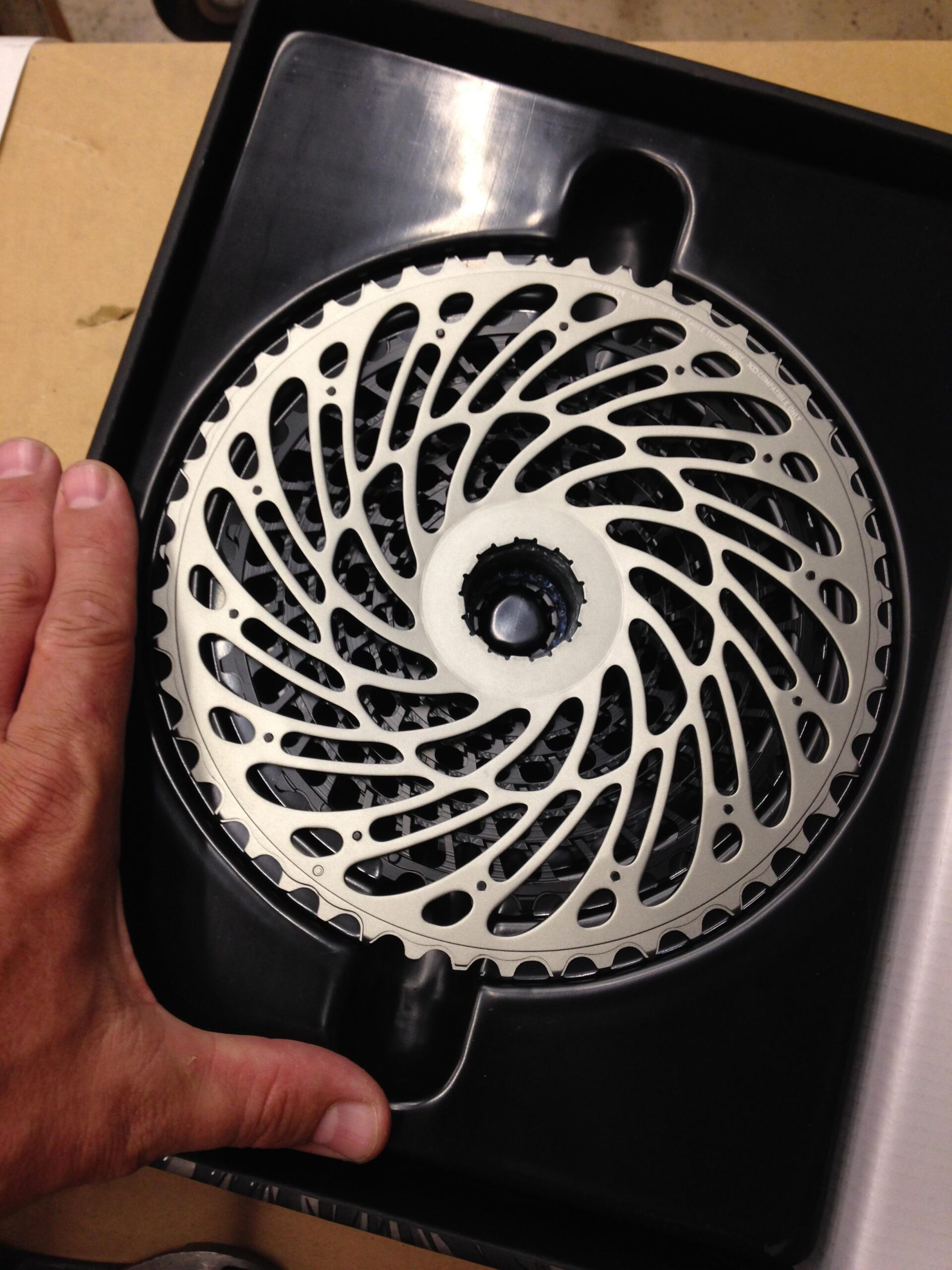 A hand holds a two-part bicycle rotor designed for disc brakes, featuring an intricate, swirling pattern. The rotor rests in a black plastic tray, highlighting its silver finish and detailed design. The background appears neutral and slightly blurred, focusing on the rotor.