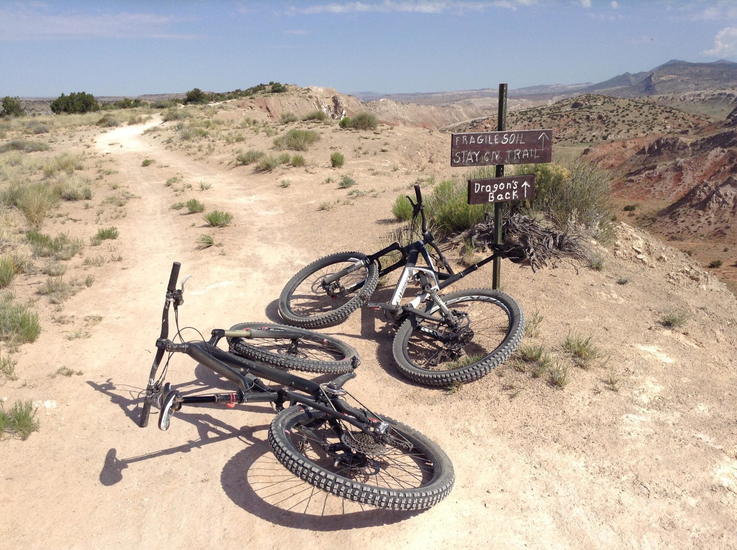 Two mountain bikes lie on a dirt trail surrounded by sparse vegetation. A signpost stands nearby, indicating "Fragile Soil" and instructing to "Stay on Trail" with directions to "Dragon's Back." The landscape features rocky terrain and rolling hills under a clear blue sky. White Ridge Bike Trails mountain bike trail.