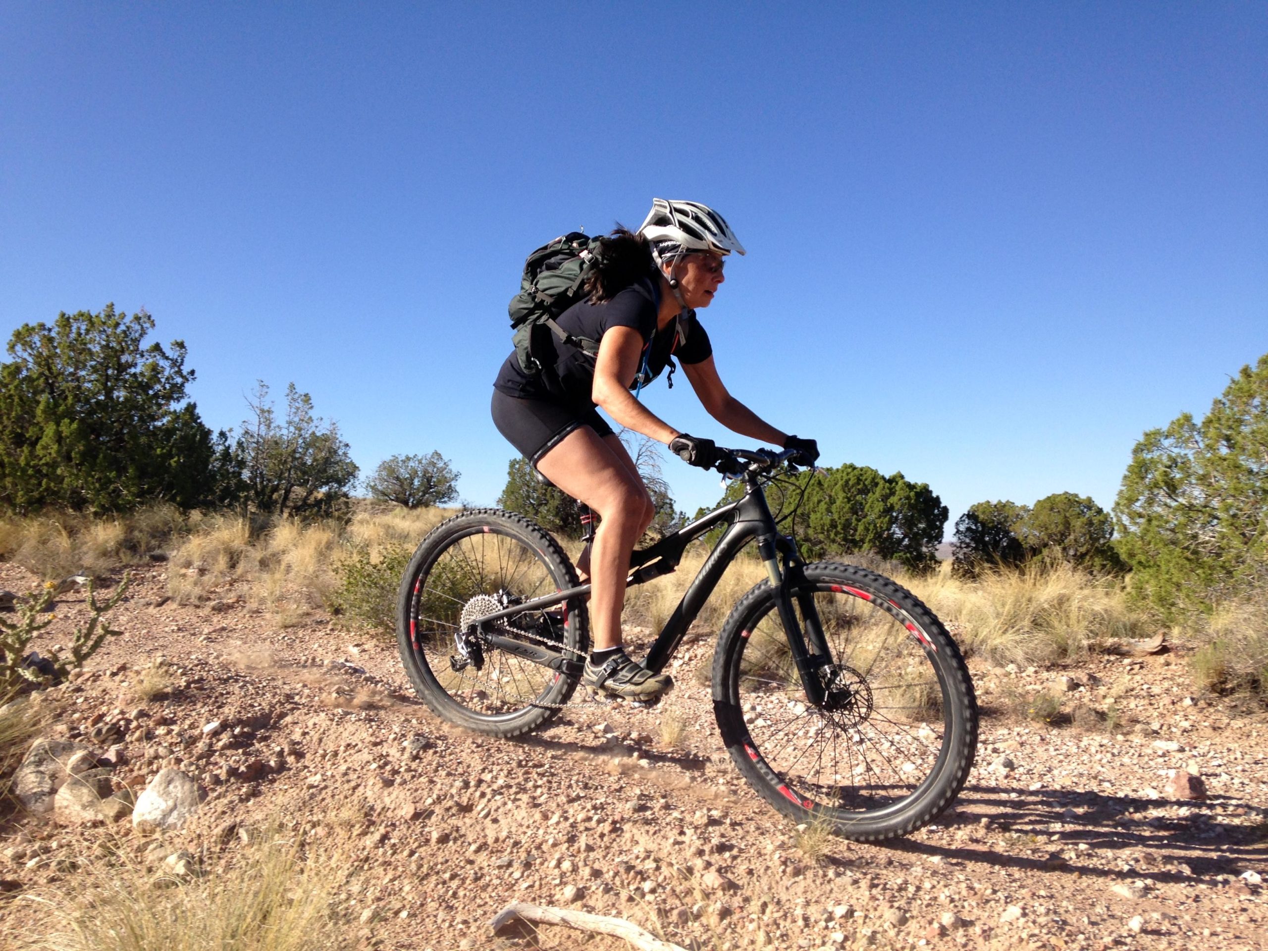 A mountain biker wearing a helmet and a backpack rides along a rocky trail in a desert-like landscape, with sparse vegetation and a clear blue sky in the background. Middle Reservoir Trail mountain bike trail.