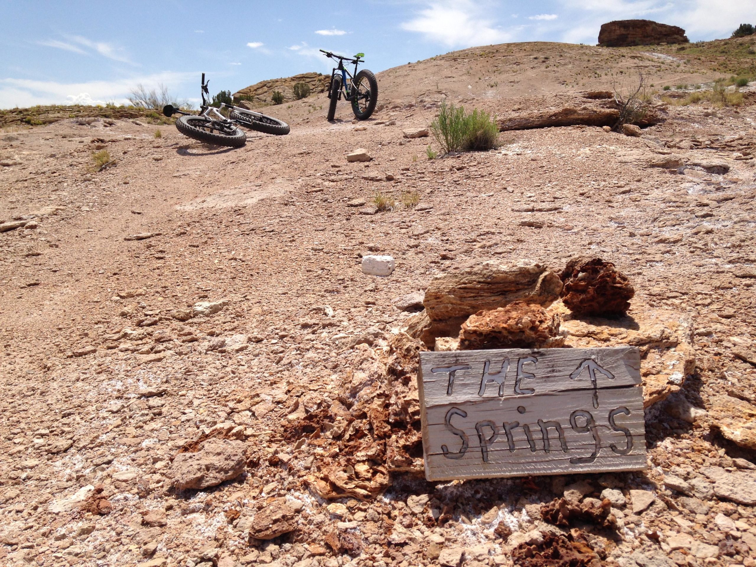 A dusty landscape with two mountain bikes parked on a rocky terrain. In the foreground, a wooden sign reads "THE Springs" with an arrow pointing upwards. The background features a hilly area with sparse vegetation and a clear blue sky. White Ridge Bike Trails mountain bike trail.