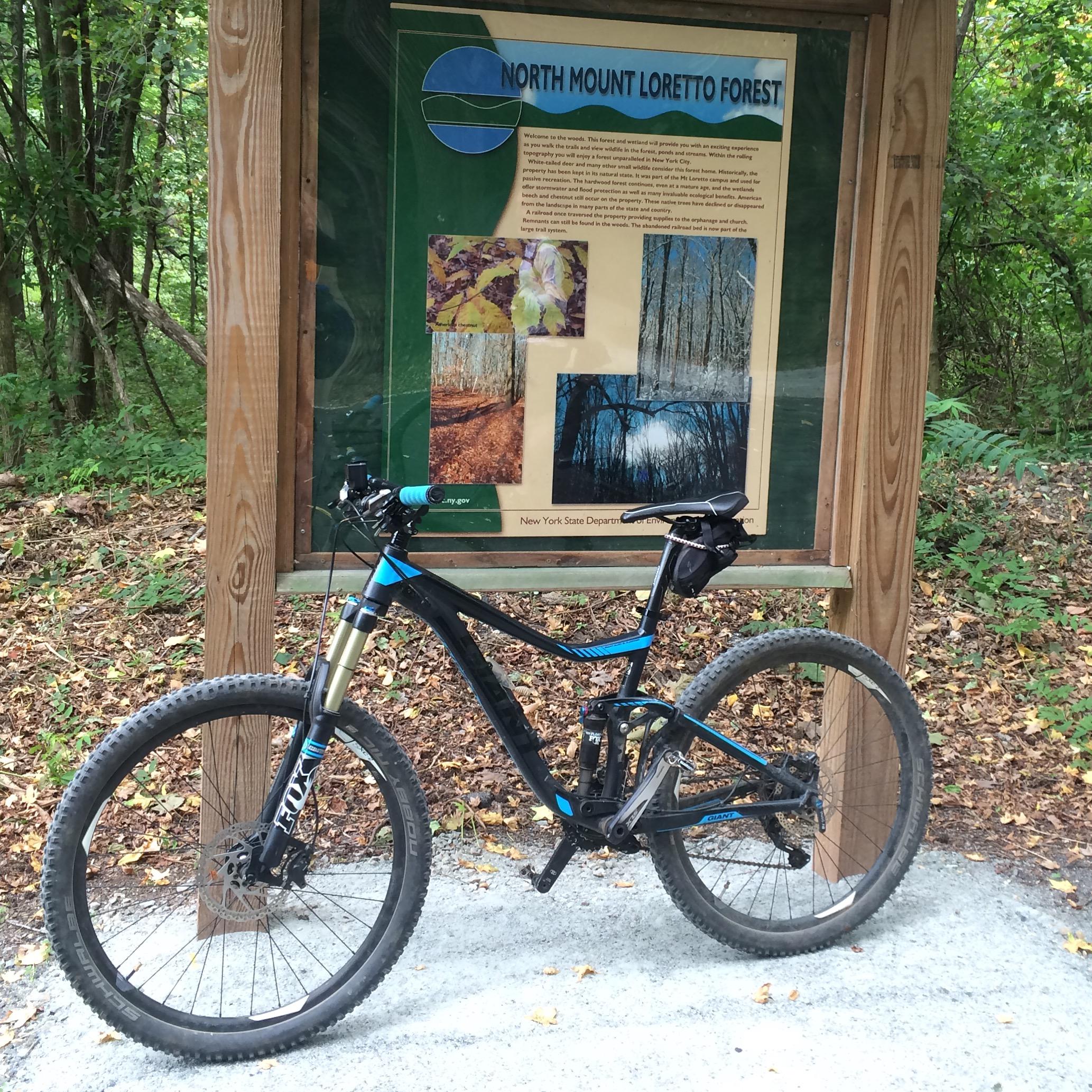 A mountain bike rests next to an informational sign about North Mount Loretto Forest, surrounded by green foliage and autumn leaves. The sign features descriptions and images of the forest's landscapes and natural features. Long Pond mountain bike trail.