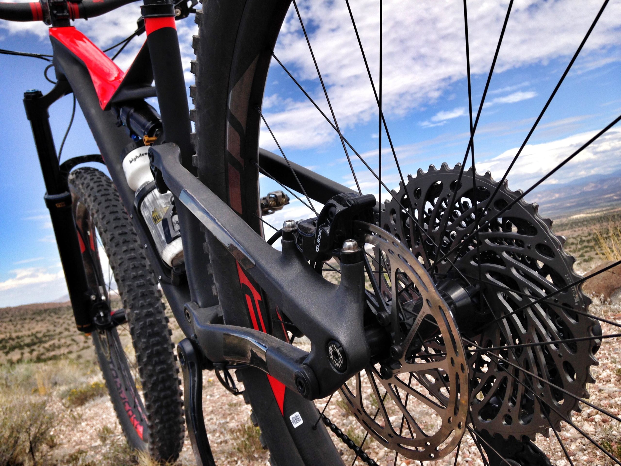 Close-up image of a mountain bike showcasing its rear wheel, brake rotor, and chainstay. The background features a scenic landscape with blue skies and distant hills. The bike has a sleek, modern design with a distinctive red and black color scheme. Mariposa Fat Bike Trails mountain bike trail.