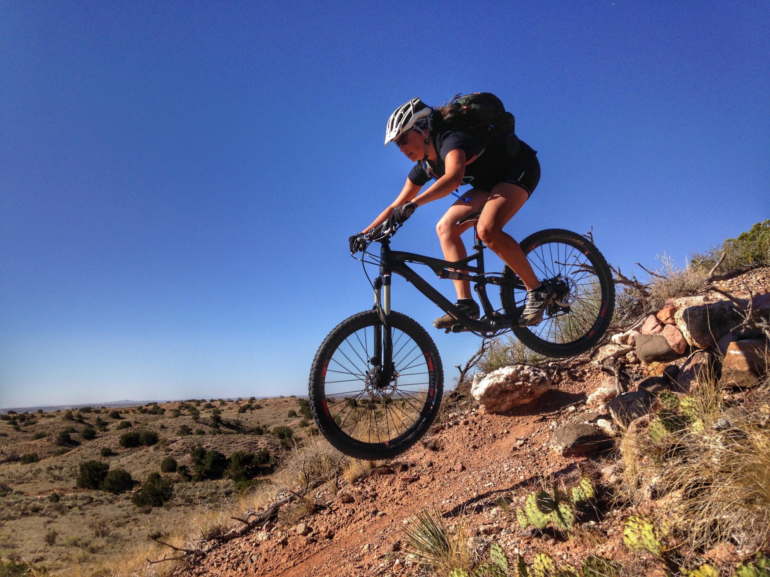A mountain biker in mid-air jumps off a rocky trail against a clear blue sky, showcasing an action shot in a rugged, outdoor landscape. The rider is wearing a helmet and backpack, demonstrating an adventurous spirit in a natural setting. Mariposa Fat Bike Trails mountain bike trail.