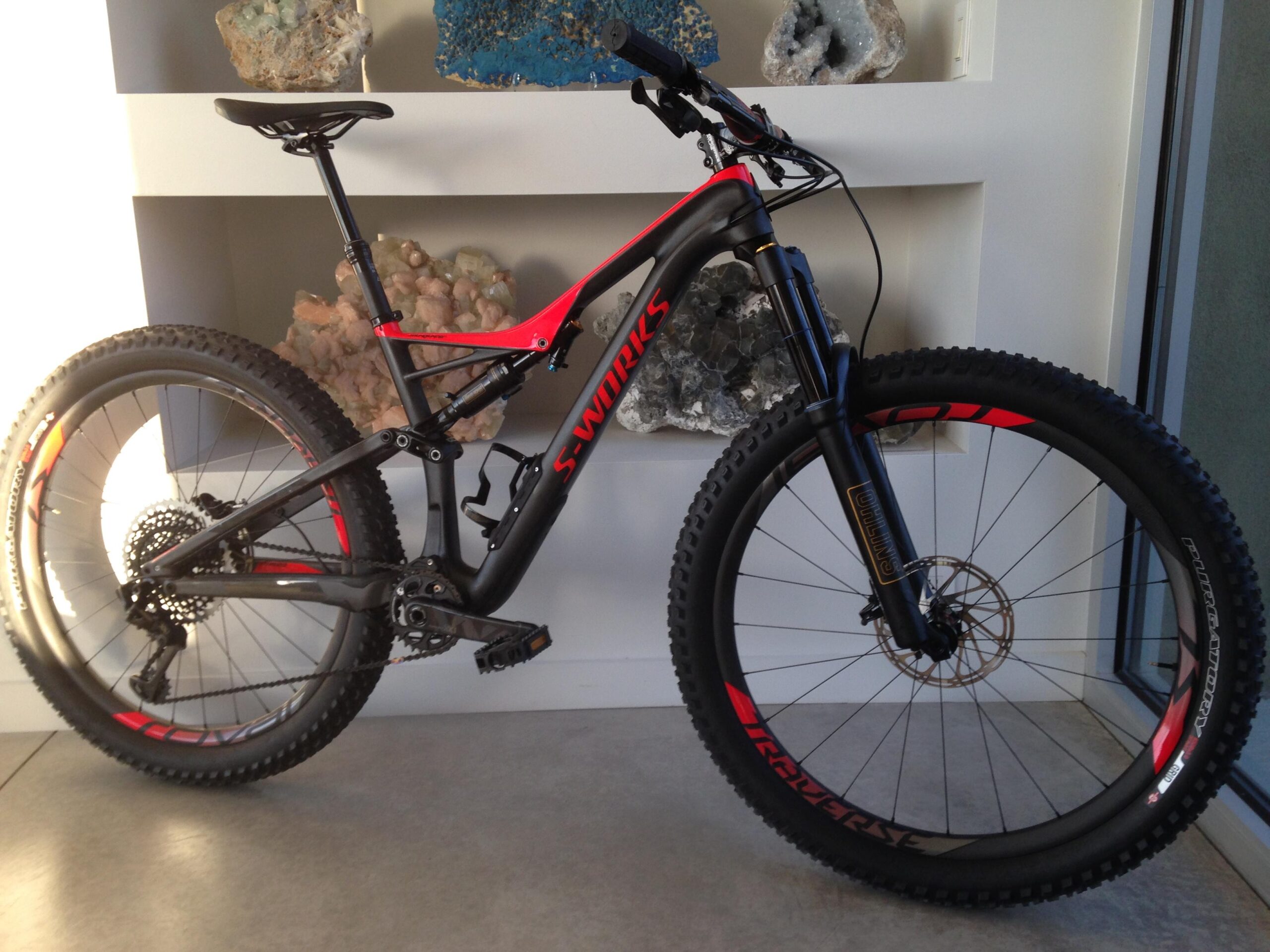 Specialized Stumpjumper FSR 6Fattie: A black and red mountain bike is showcased against a backdrop of various rocks and crystals. The bike features a sleek design, prominent suspension system, and wide tires suitable for off-road riding. It is positioned upright on a modern concrete floor.