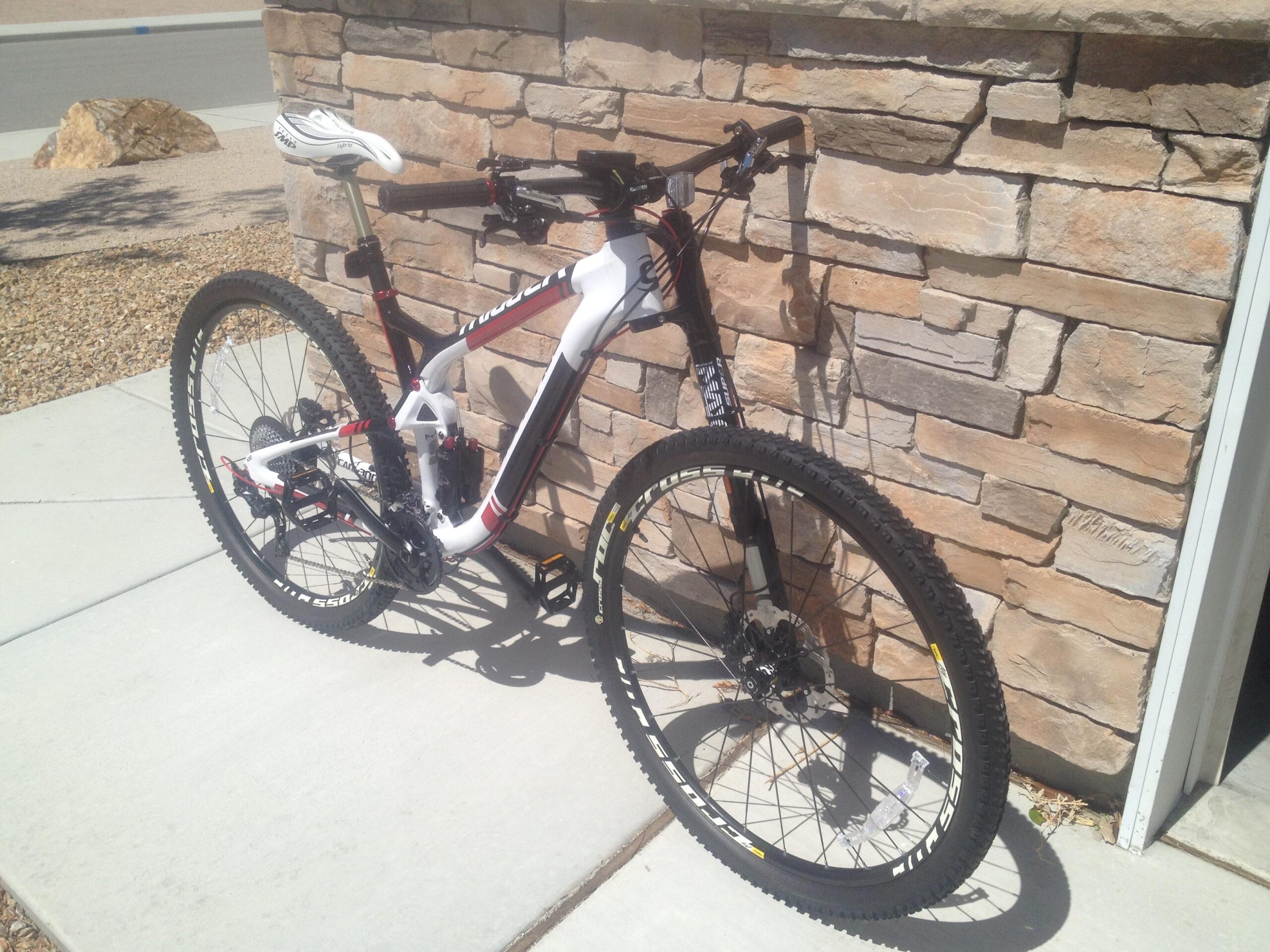 Cannondale Trigger 29 Carbon 1: A modern mountain bike leaning against a stone wall, featuring a white and red frame, knobby tires, and various components visible, situated on a concrete pathway with a gravel area nearby.