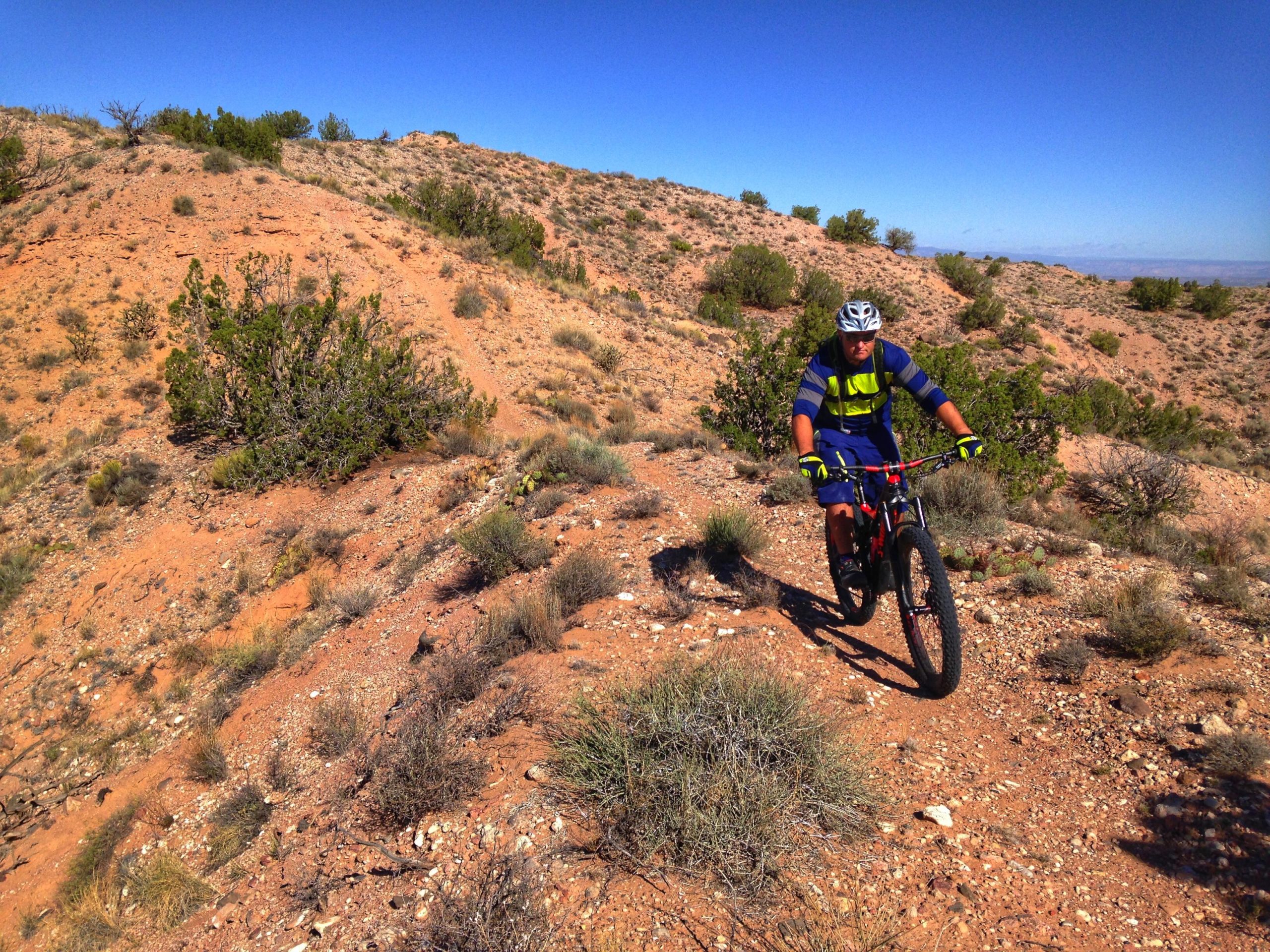 A mountain biker riding along a rocky trail in a desert landscape, surrounded by sparse vegetation and hills under a clear blue sky. The cyclist is wearing a helmet, bright clothing, and gloves, and appears focused on navigating the trail. Mariposa Fat Bike Trails mountain bike trail.