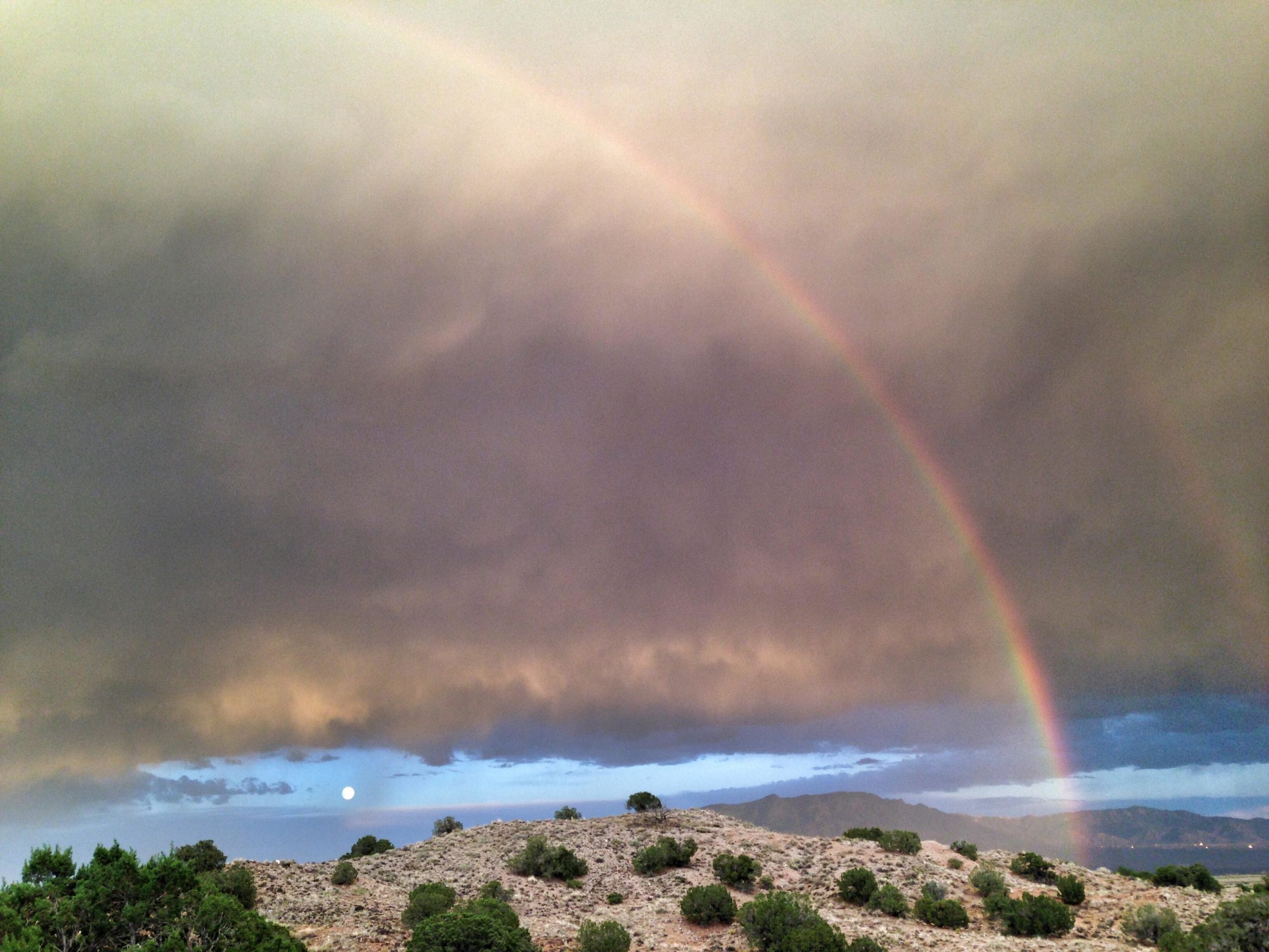 A beautiful landscape featuring a vibrant rainbow arching across a cloudy sky, with a pale moon visible on the horizon. The foreground includes rocky terrain and sparse vegetation, while distant mountains are seen beneath the colorful sky. Mariposa Fat Bike Trails mountain bike trail.