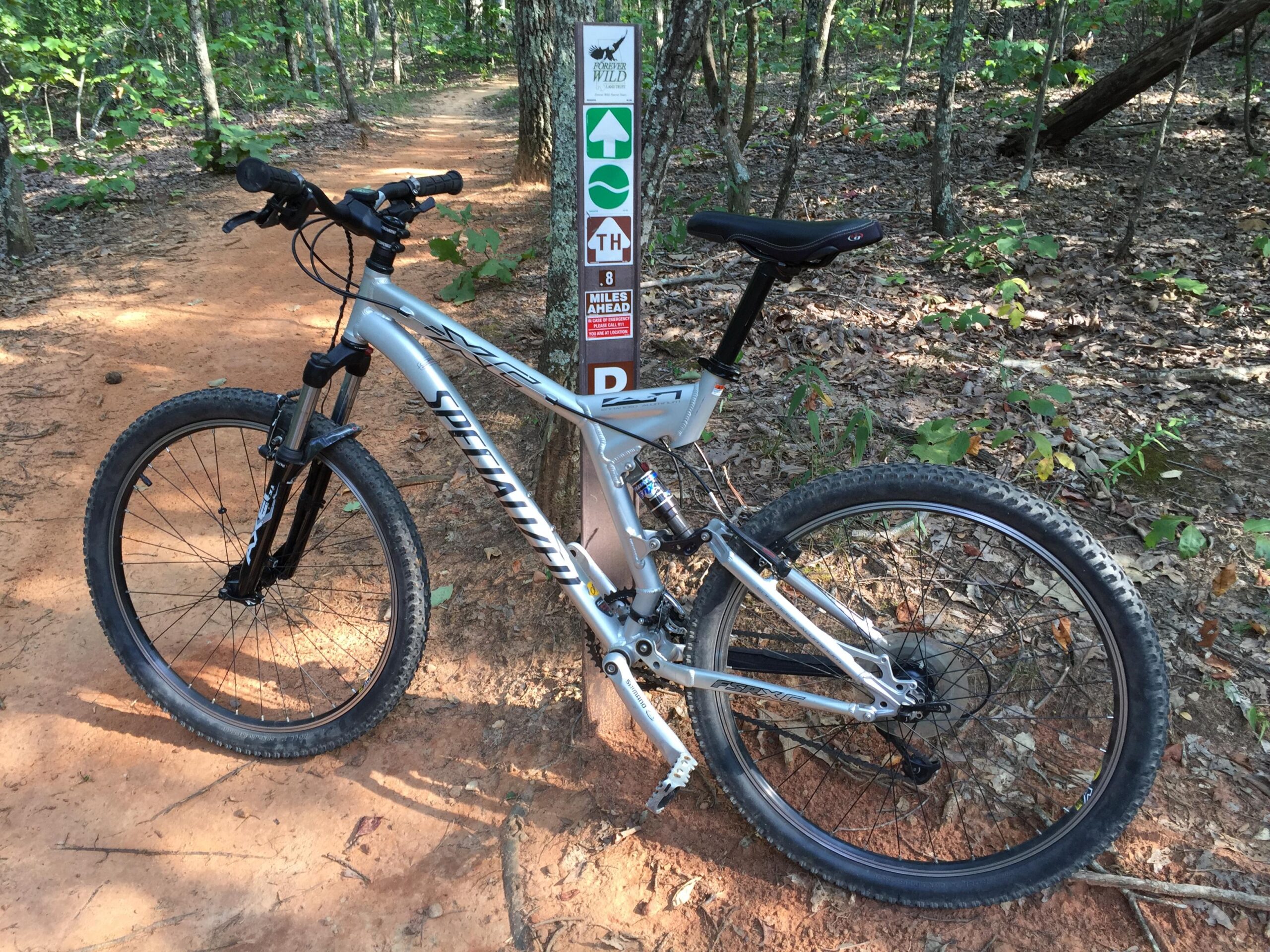 Specialized FSRxc Pro: Mountain bike resting on a dirt trail surrounded by trees, with a trail sign indicating directions and distance ahead.
