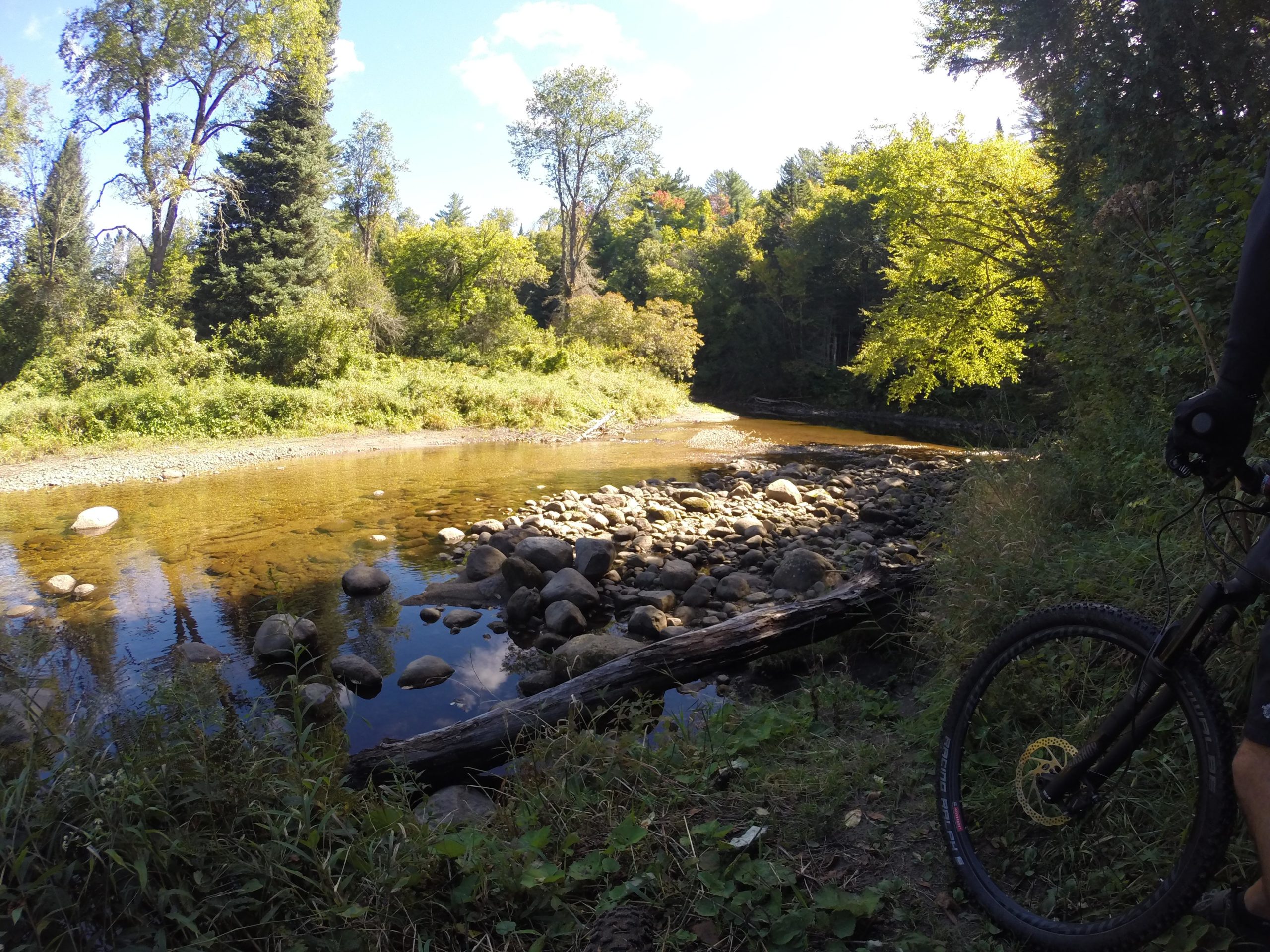 A scenic view of a calm river surrounded by lush greenery, featuring large rocks in the water and along the riverbank. Sunlight filters through the trees, creating a serene atmosphere, with a mountain bike partially visible in the foreground. Kingdom Trails mountain bike trail.