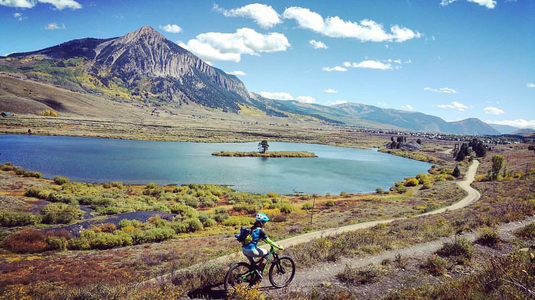 A person riding a mountain bike on a dirt path beside a serene lake surrounded by autumn-colored vegetation, with a large mountain in the background under a partially cloudy blue sky. Budd Trail mountain bike trail.