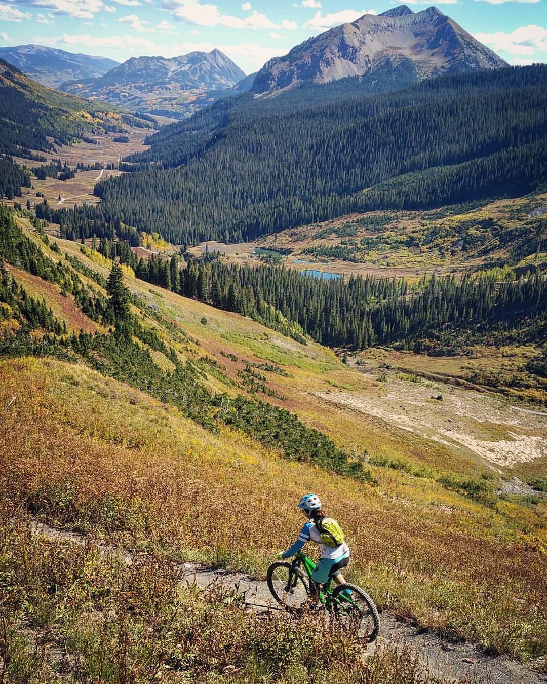 A mountain biker navigates a winding trail through a lush, mountainous landscape, surrounded by a mix of green forests and colorful autumn foliage. Towering peaks rise in the background under a clear blue sky, while a serene valley is visible below. Trail 401 mountain bike trail.