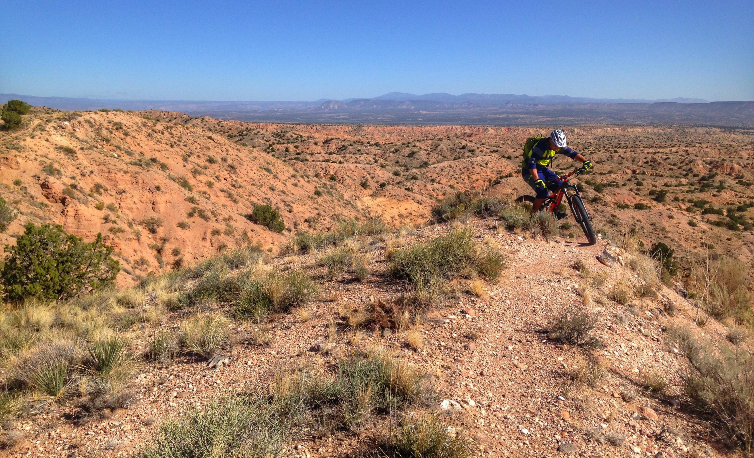 A mountain biker navigating a rugged trail on a hillside with a vast desert landscape and distant mountains in the background, under a clear blue sky. The terrain features rocky soil and sparse vegetation. Mariposa Fat Bike Trails mountain bike trail.