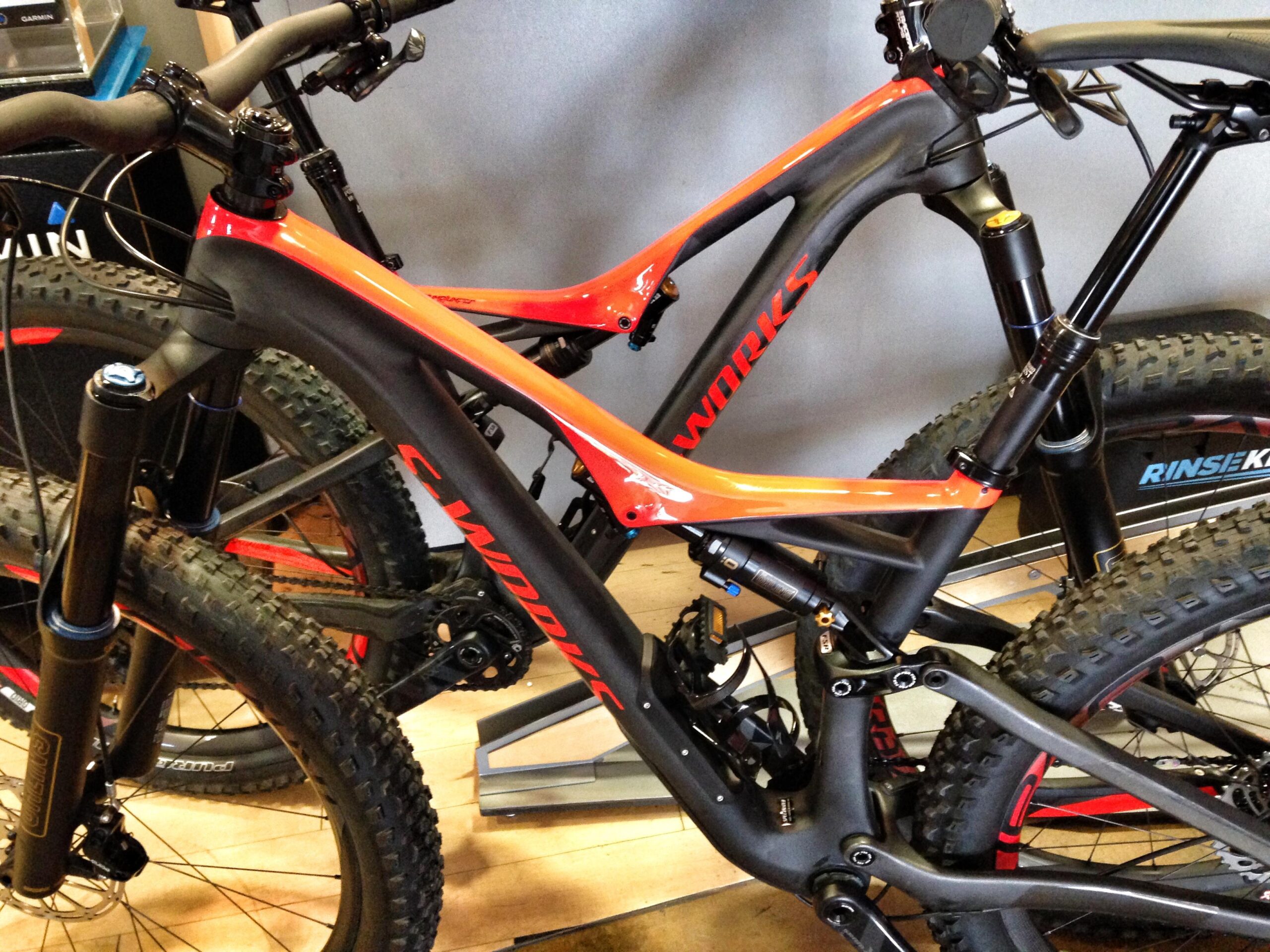 Close-up image of two mountain bikes showcasing a solid black and orange frame design, featuring intricate components like the fork, handlebars, and drive train. The bikes are displayed in a retail setting with a wooden floor, highlighting their rugged tires and advanced suspension systems.