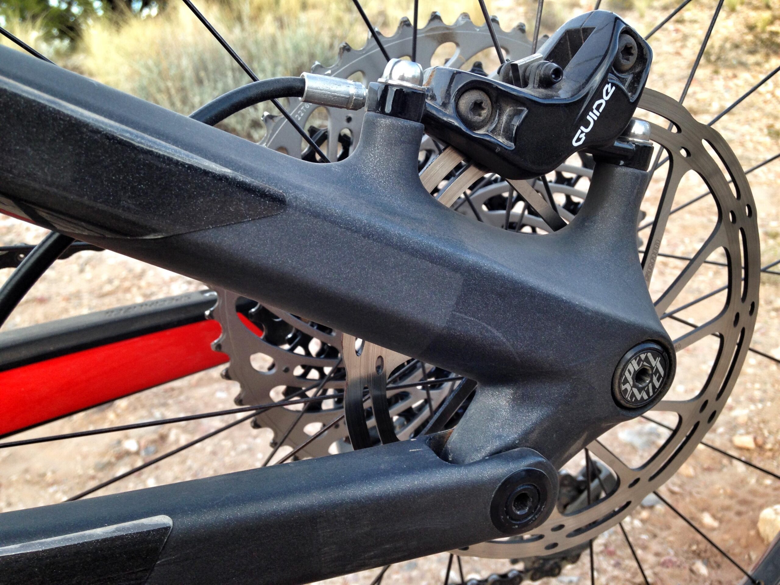 Specialized Stumpjumper FSR 6Fattie: A close-up view of a bicycle's rear brake assembly, featuring a black brake caliper labeled "Guide" attached to the frame. The image highlights the intricate design of the frame, a red tube, and the circular metal disc brake, set against a blurred natural background.