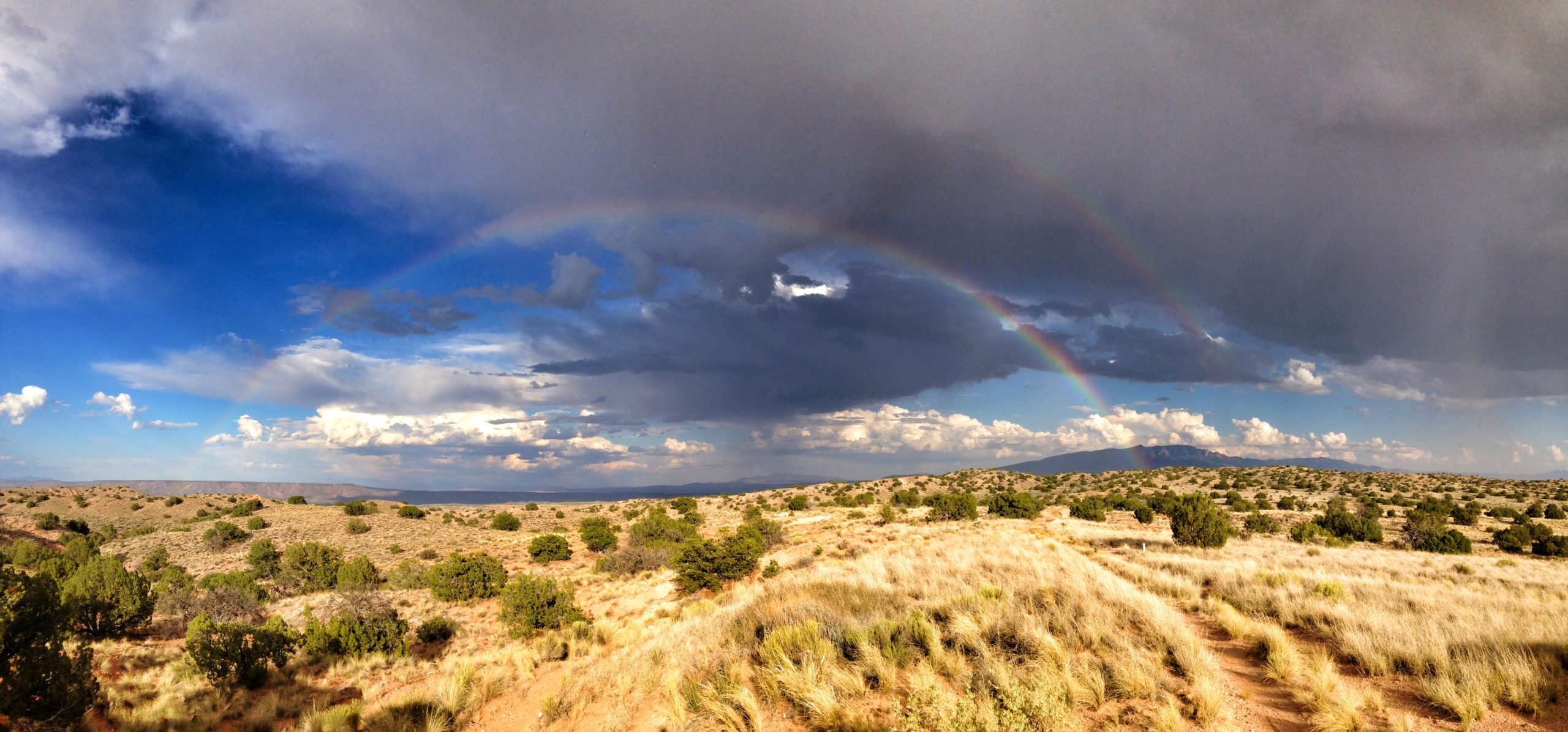 Panoramic view of a desert landscape with rolling hills and sparse vegetation, featuring a vibrant rainbow arching across a dramatic sky filled with dark clouds and patches of blue. The foreground includes dry grasses and a winding dirt path leading into the scene. Mariposa Fat Bike Trails mountain bike trail.