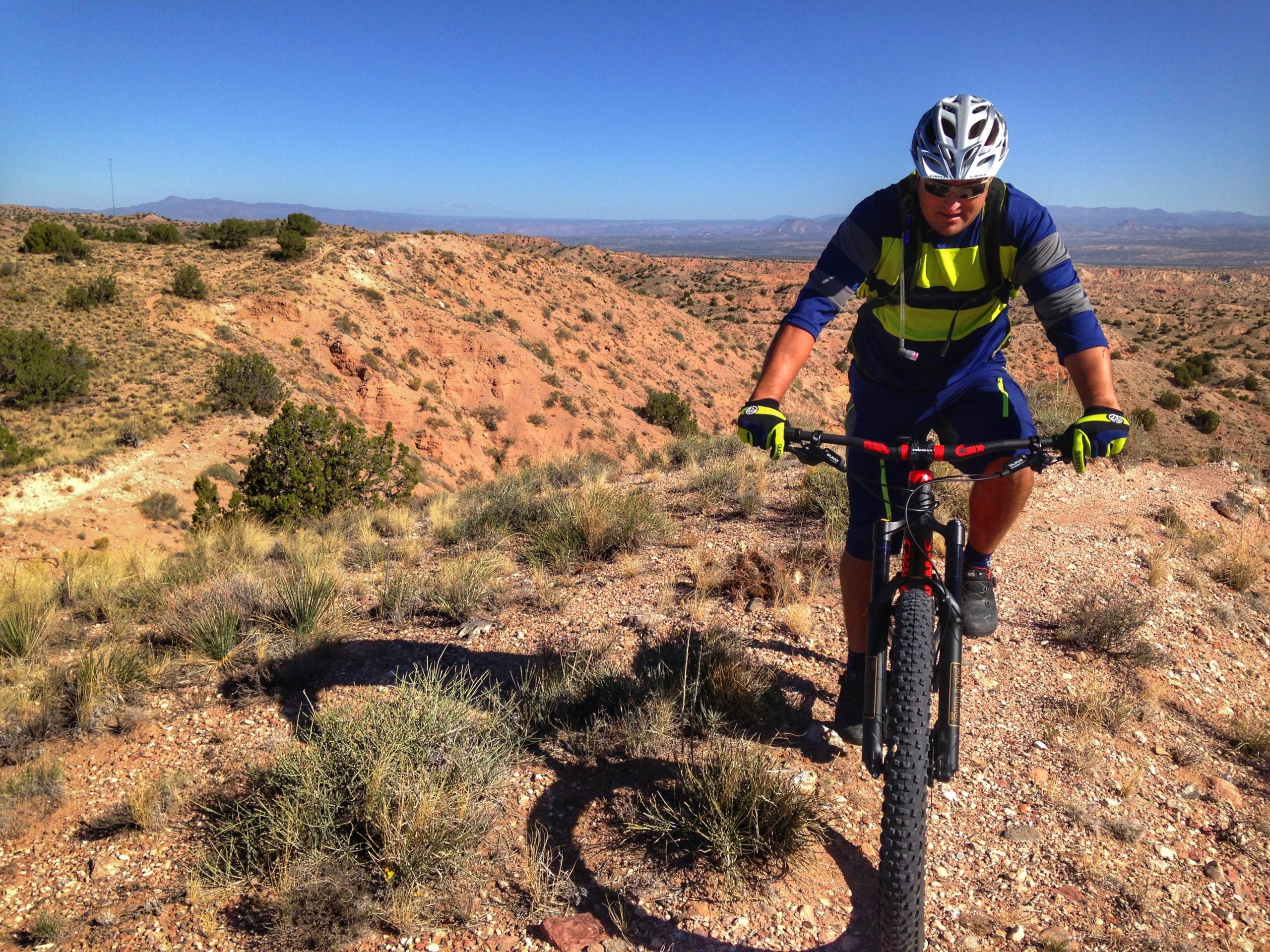 A man in a bright blue and yellow cycling outfit rides a mountain bike on a rocky trail surrounded by desert landscapes. The sun is shining against a clear blue sky, highlighting the rugged terrain and sparse vegetation in the background. Mariposa Fat Bike Trails mountain bike trail.