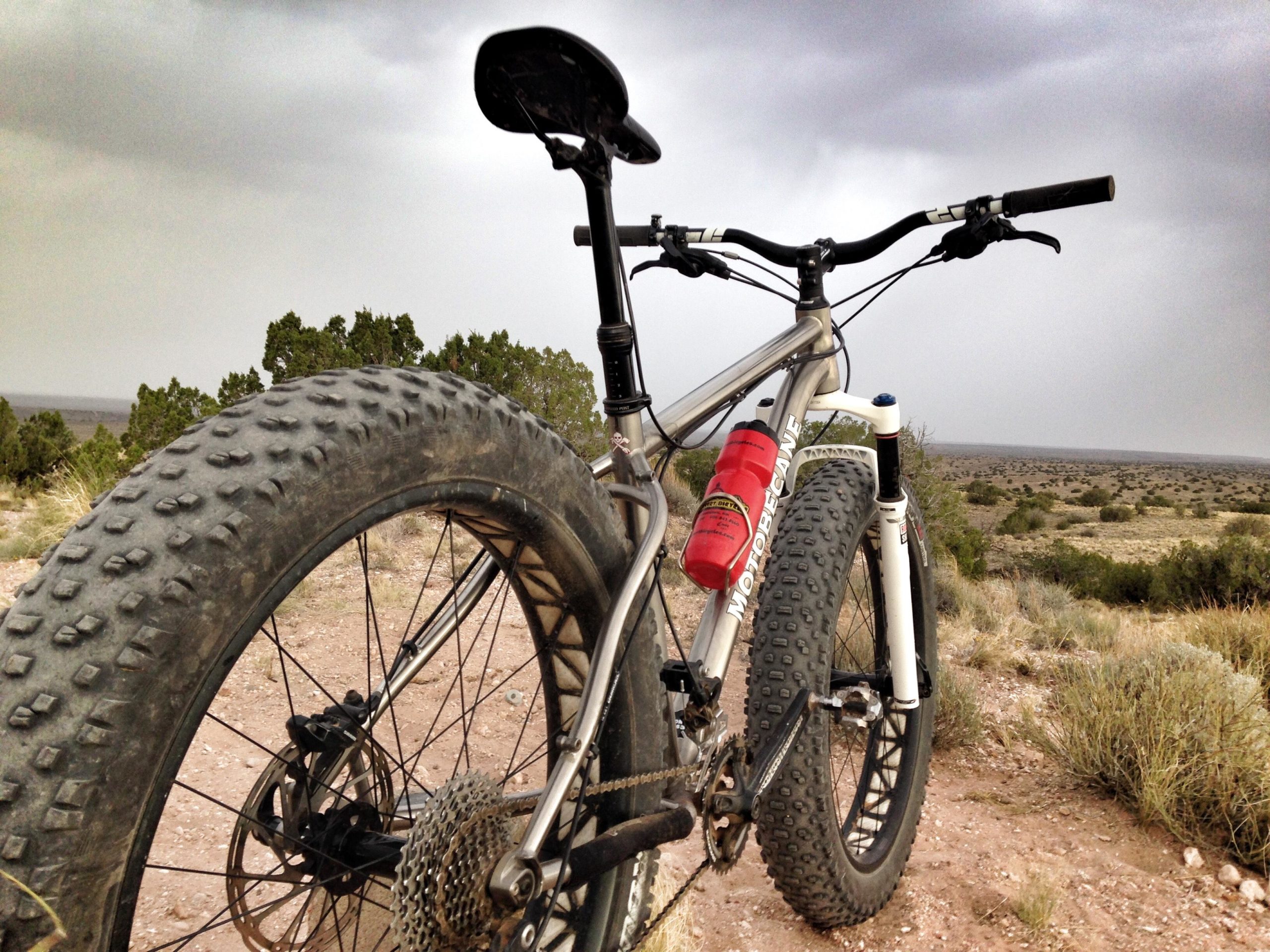 A fat tire mountain bike parked on a dirt path, with a dramatic sky in the background. The bike features a silver frame, thick tires, and a red water bottle attached to the side. In the distance, a landscape of sparse vegetation extends under an overcast sky. Mariposa Fat Bike Trails mountain bike trail.