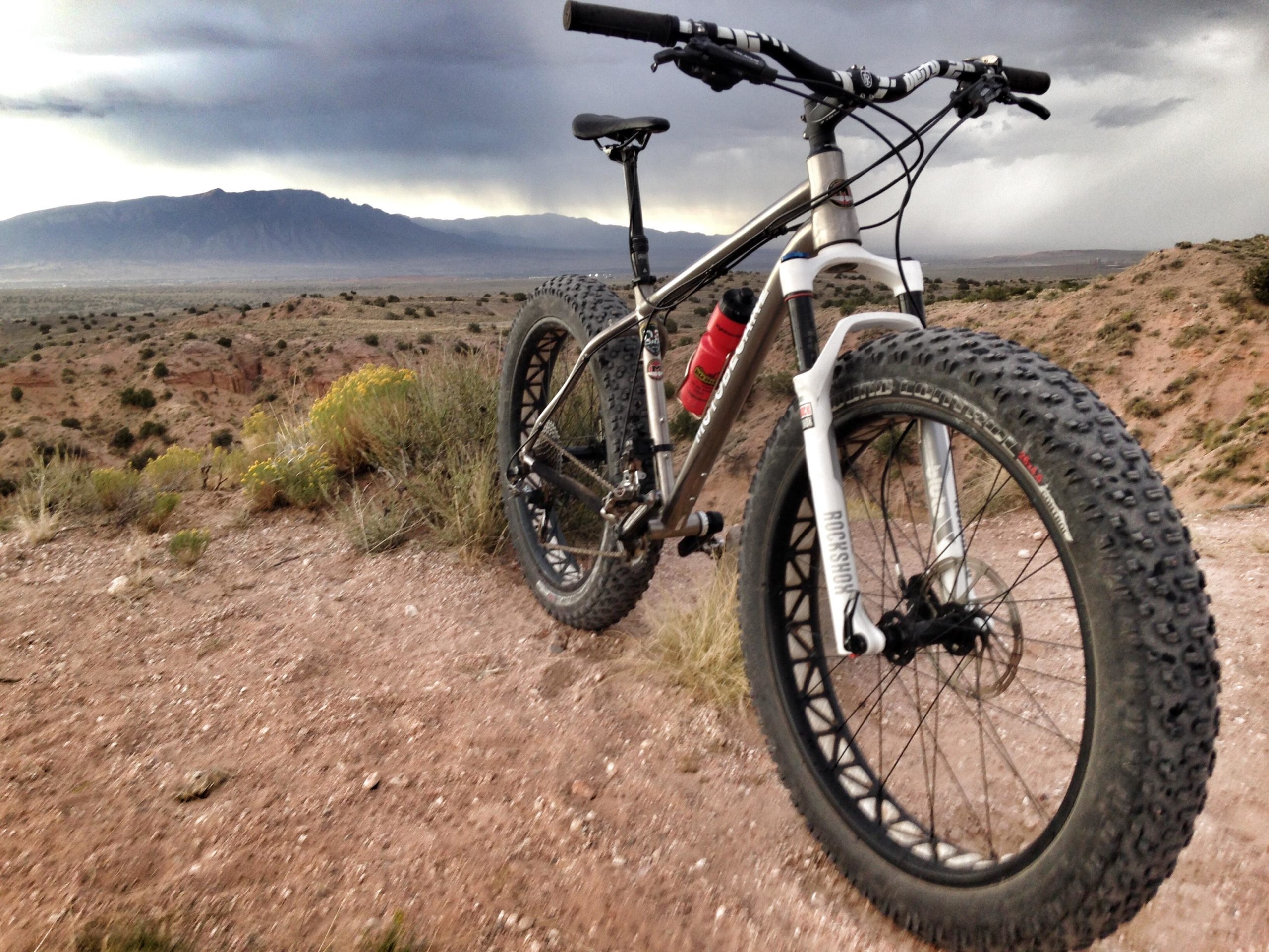 A mountain bike with thick tires is parked on a gravel trail, with a vast landscape of mountains and desert vegetation in the background. Dark clouds gather overhead, suggesting an approaching storm. Mariposa Fat Bike Trails mountain bike trail.