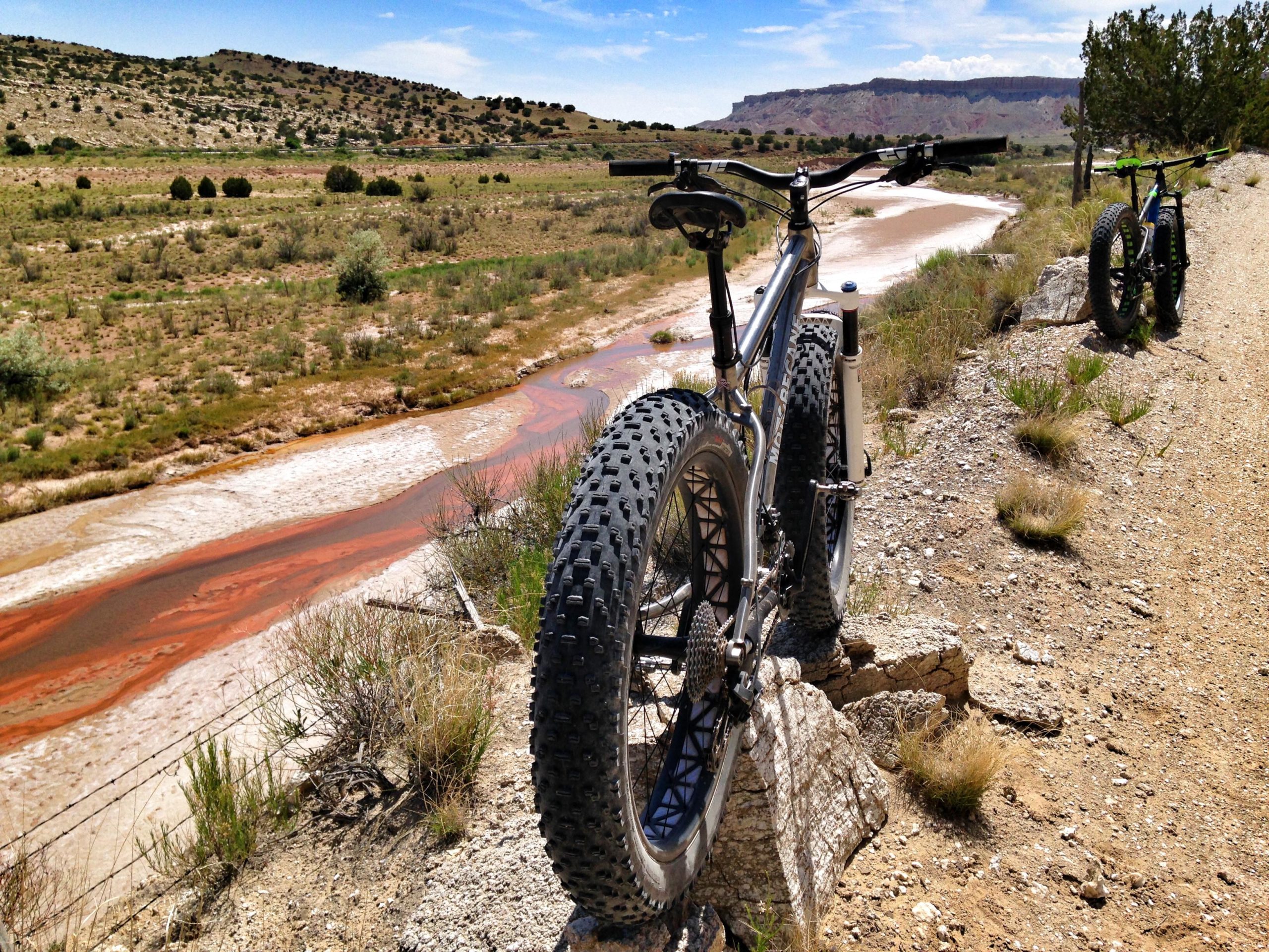 Two mountain bikes with wide tires are parked along a dirt path, overlooking a meandering river that has reddish hues. The landscape features rolling hills, sparse vegetation, and distant cliffs under a blue sky with scattered clouds. The scene captures a beautiful outdoor adventure in a rugged natural setting. White Ridge Bike Trails mountain bike trail.