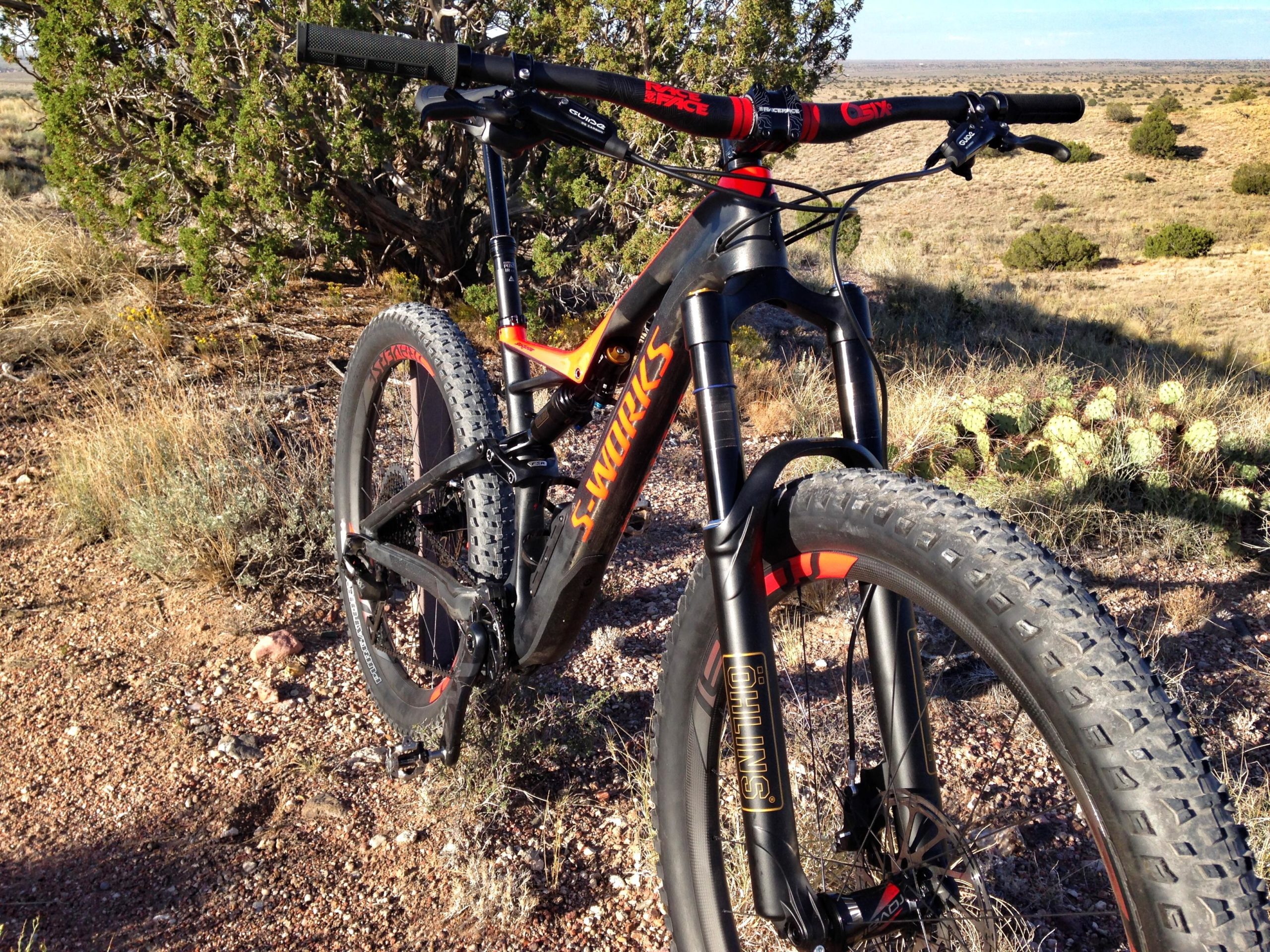 A close-up view of a mountain bike with a black and orange frame, set against a natural landscape featuring shrubs and desert vegetation. The bike is positioned on a rocky trail with visible tire treads, capturing a sense of outdoor adventure. Mariposa Fat Bike Trails mountain bike trail.